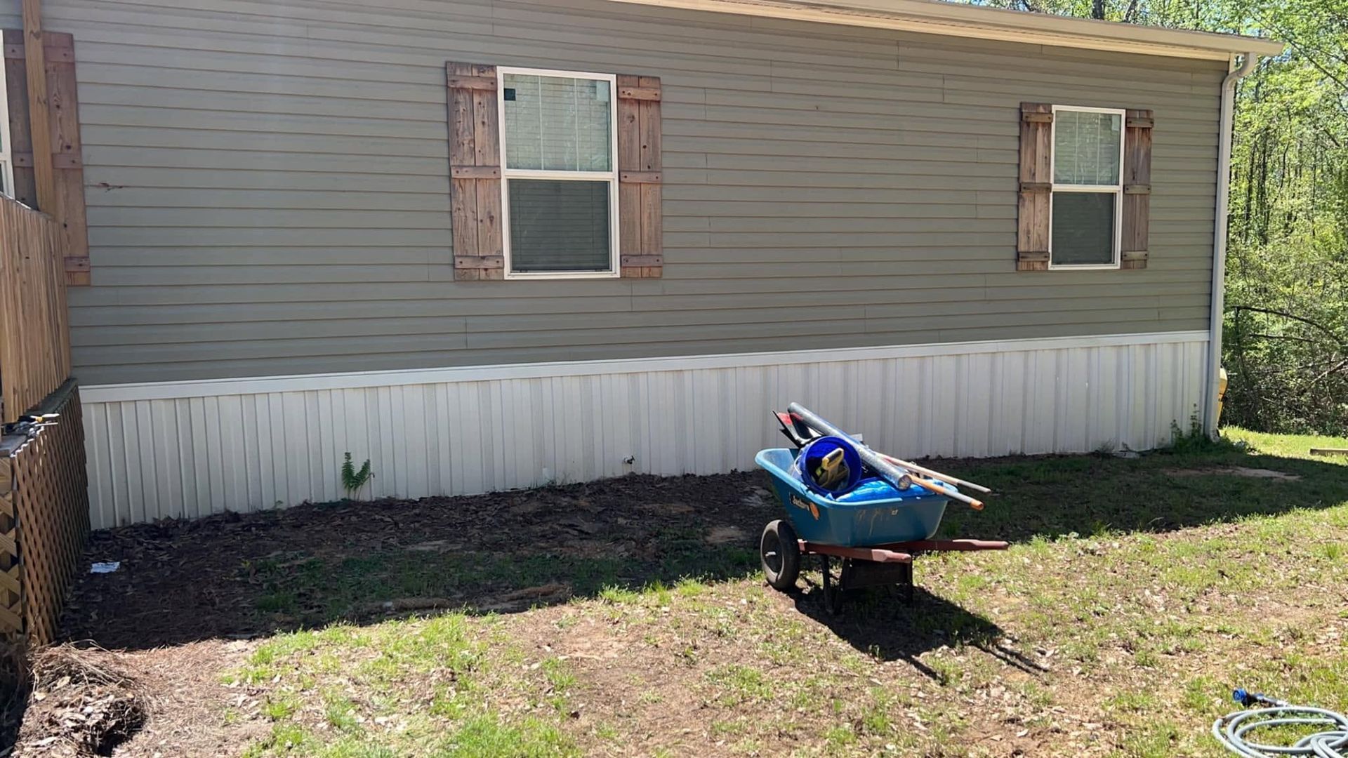 A beige mobile home with two windows and a blue wheelbarrow on the grass in front.