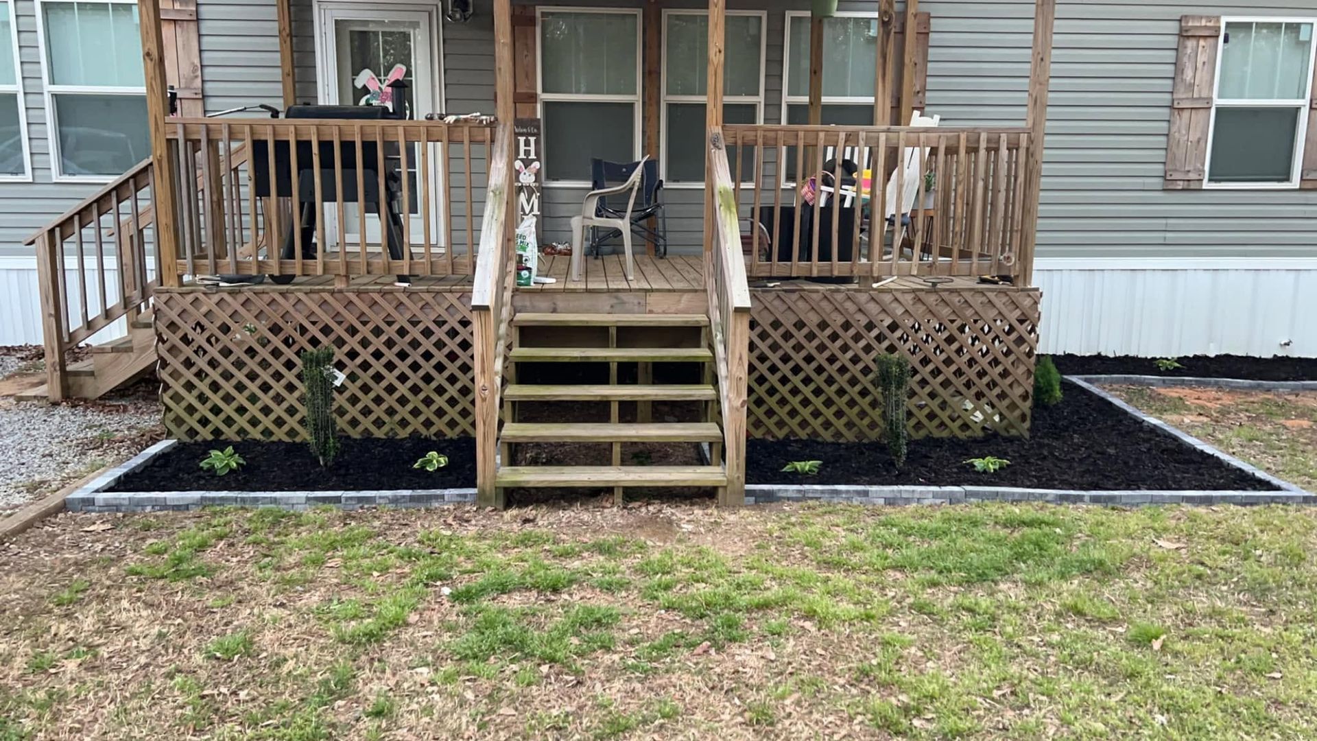 Front porch of a house with wooden steps, railing, and black mulch beds along the front yard.
