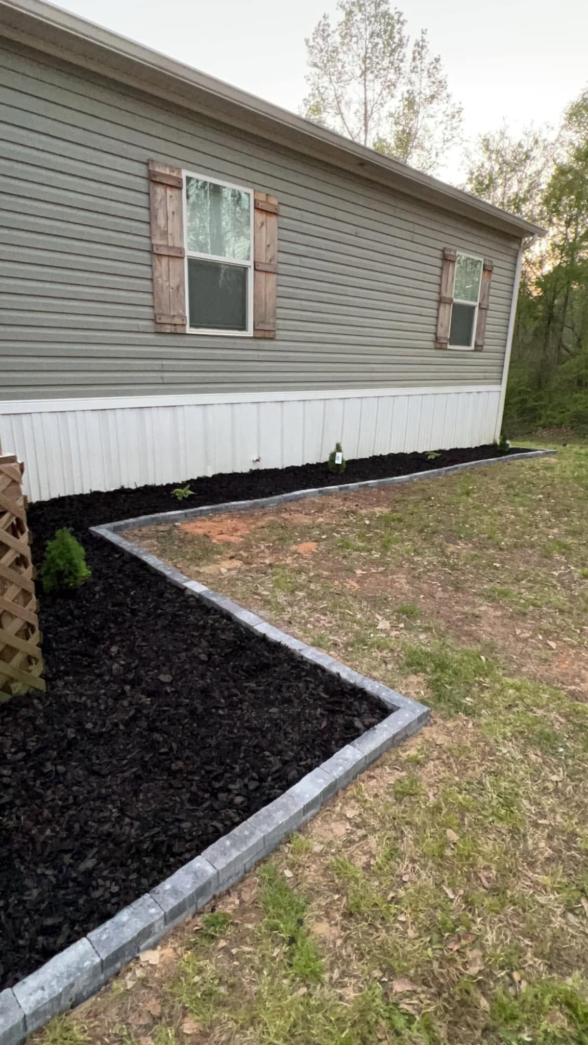 Mobile home exterior with gray siding, white trim, and a black mulch garden bed along the foundation.