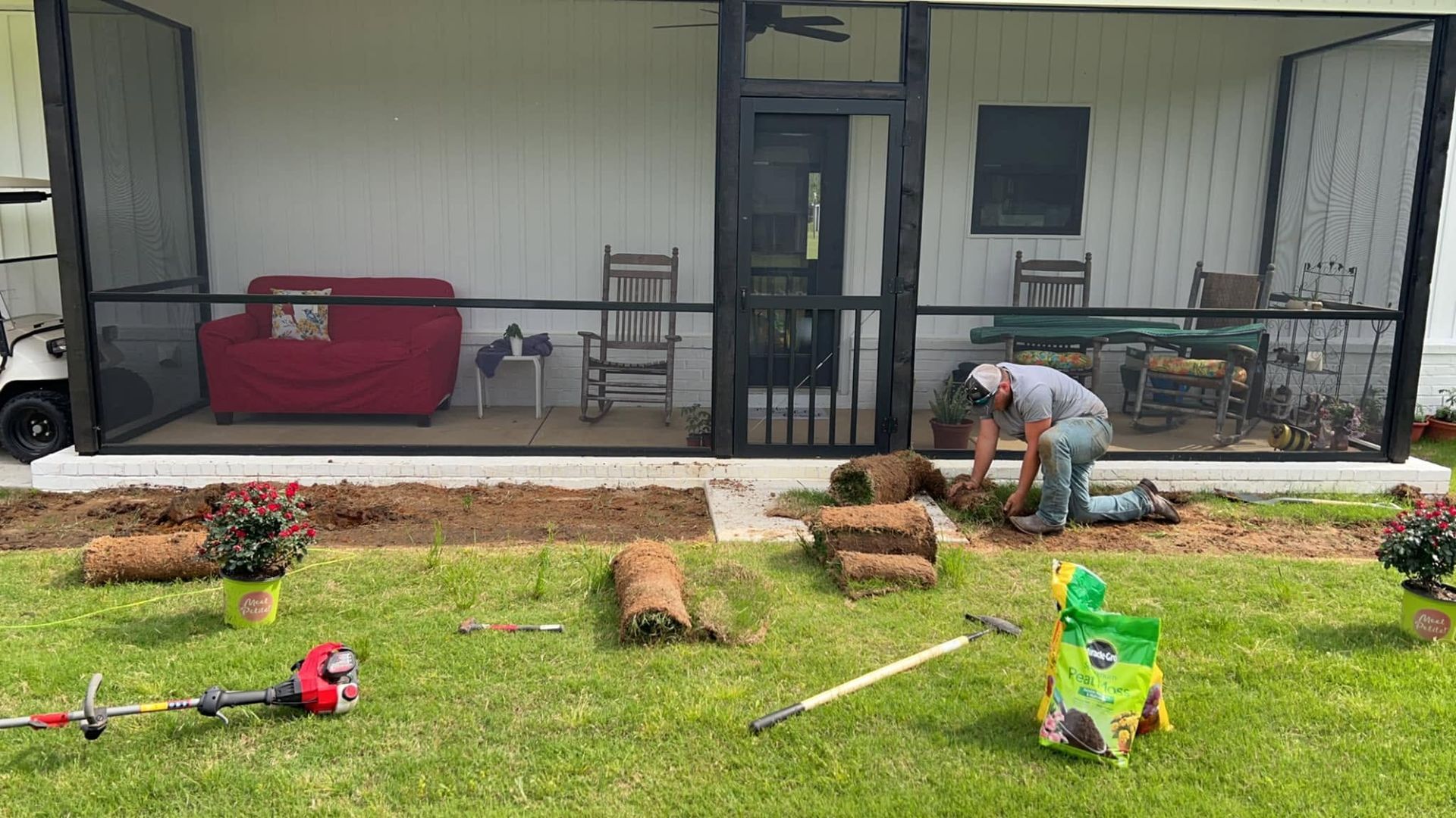 Person mowing a lawn in front of a house with scattered logs and yard tools