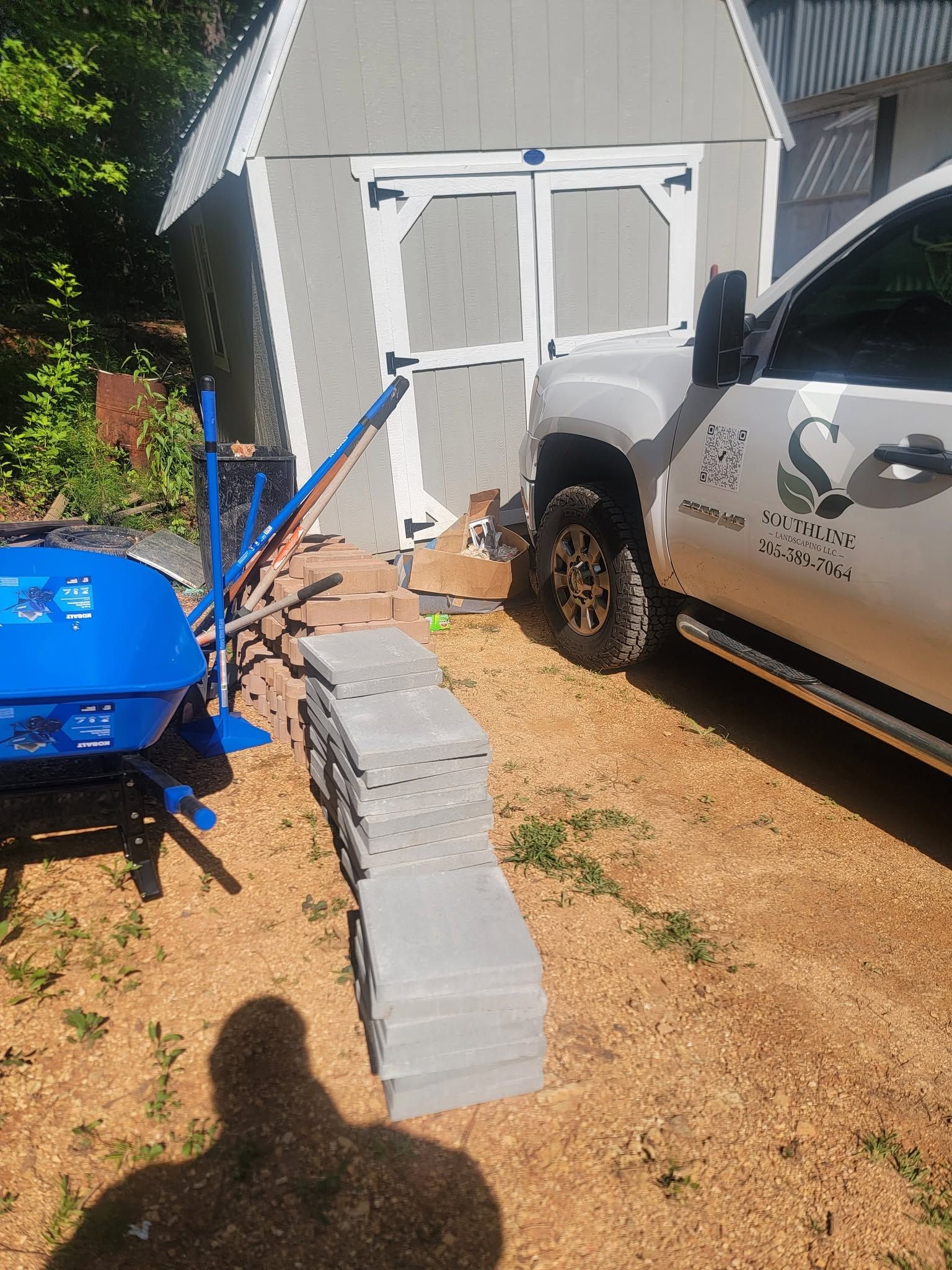 Stacked cinder blocks form a narrow path beside a white truck and blue wheelbarrow leading to a shed.
