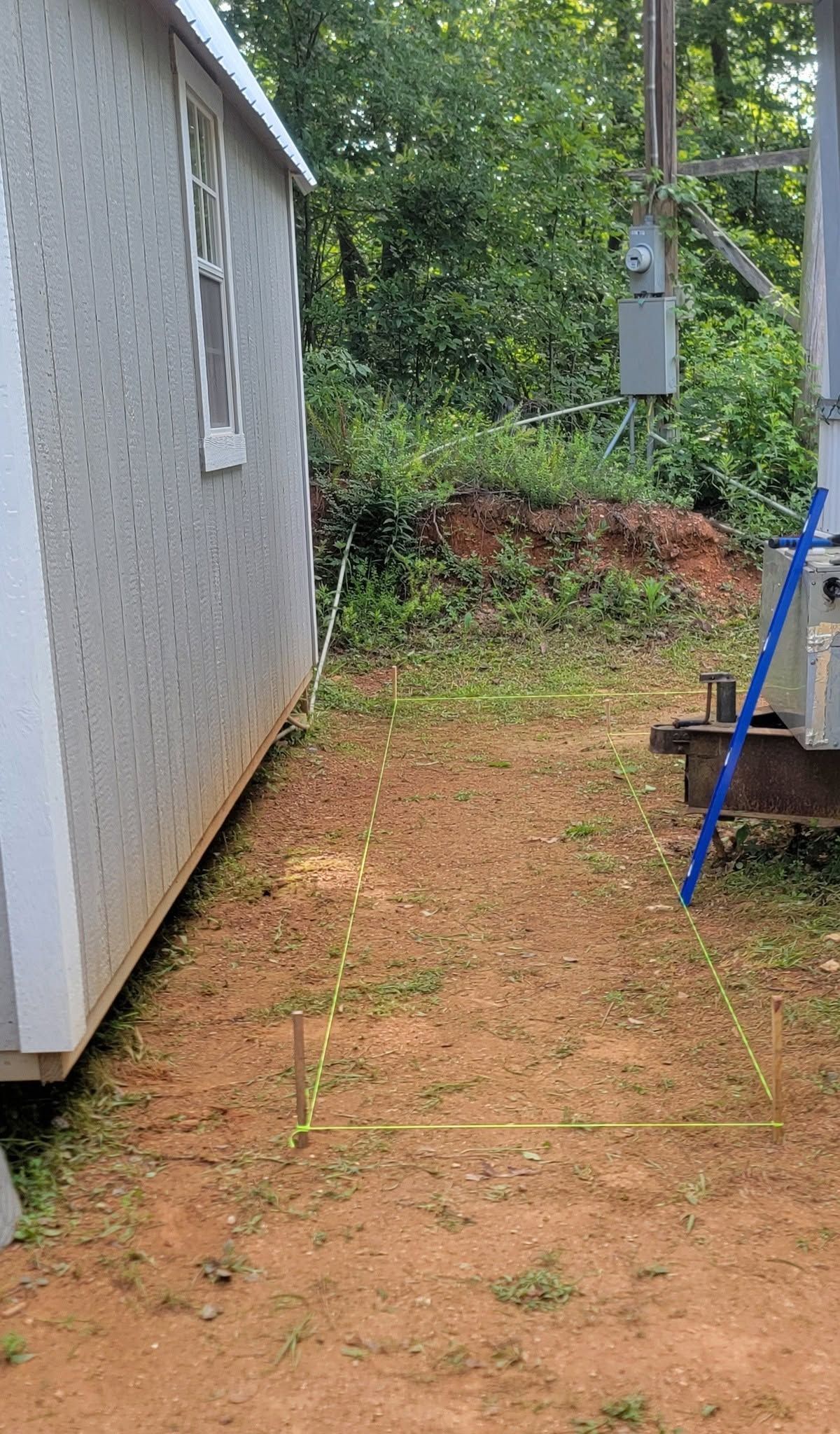Narrow dirt path beside a gray house, with green shrubs, an electrical box, and a blue ladder at right.