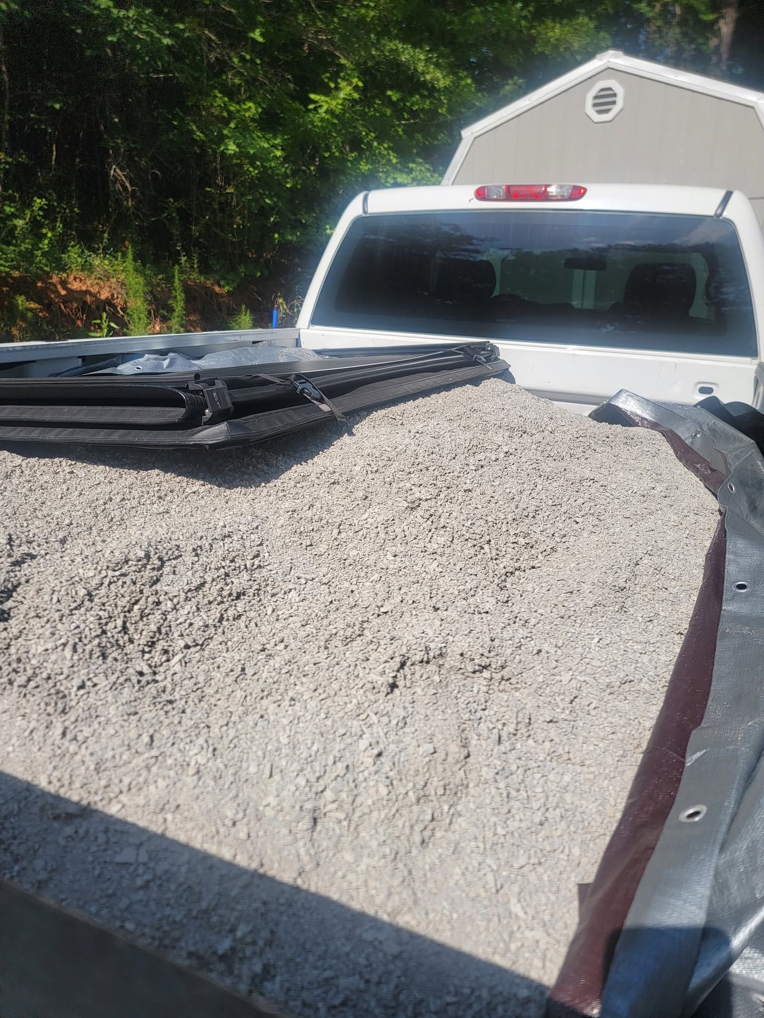 White pickup truck carrying a large gravel load in a driveway near a house and trees.