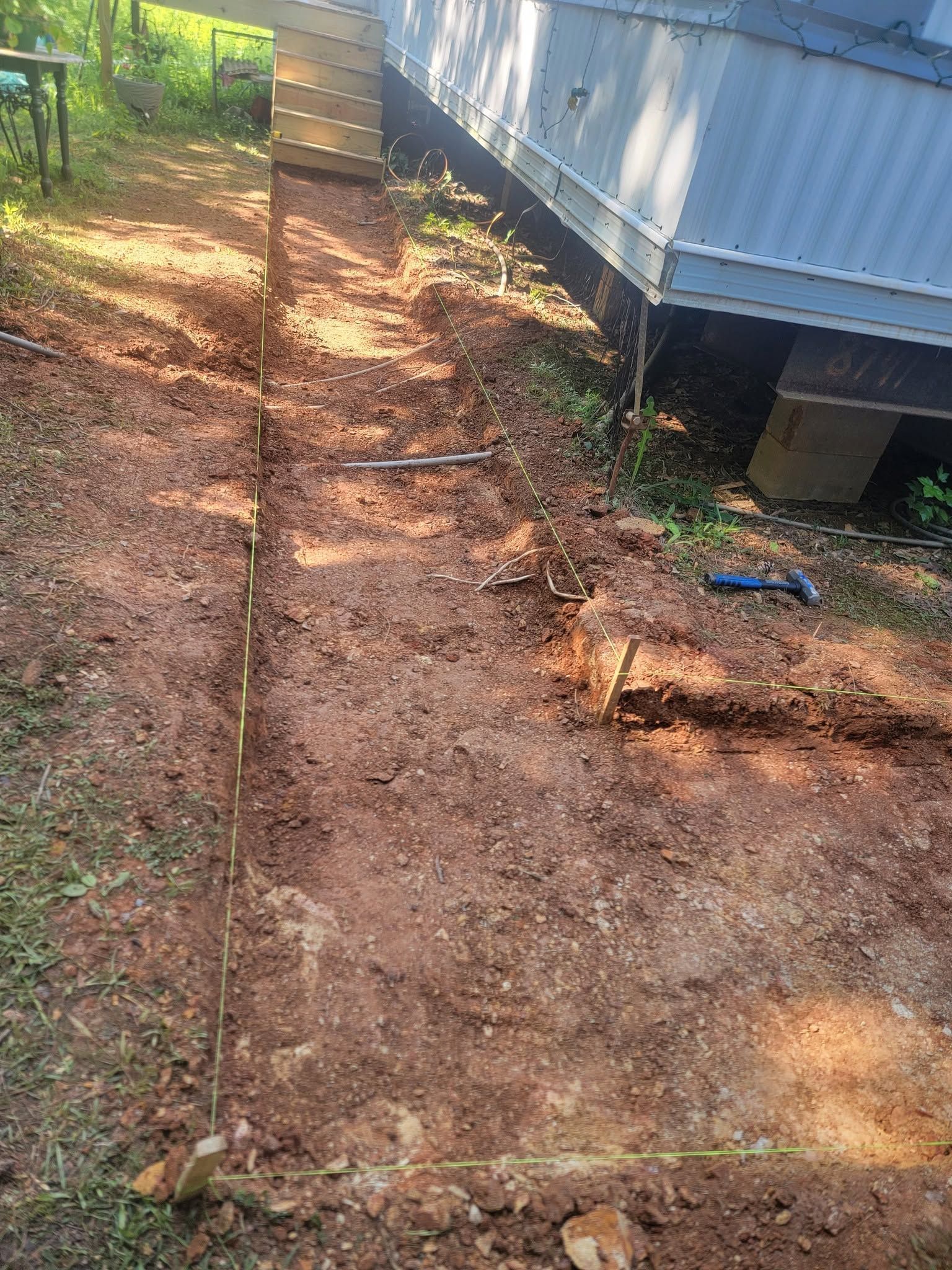 Dirt path beside a mobile home, lined with stones and bordered by trees and brush.