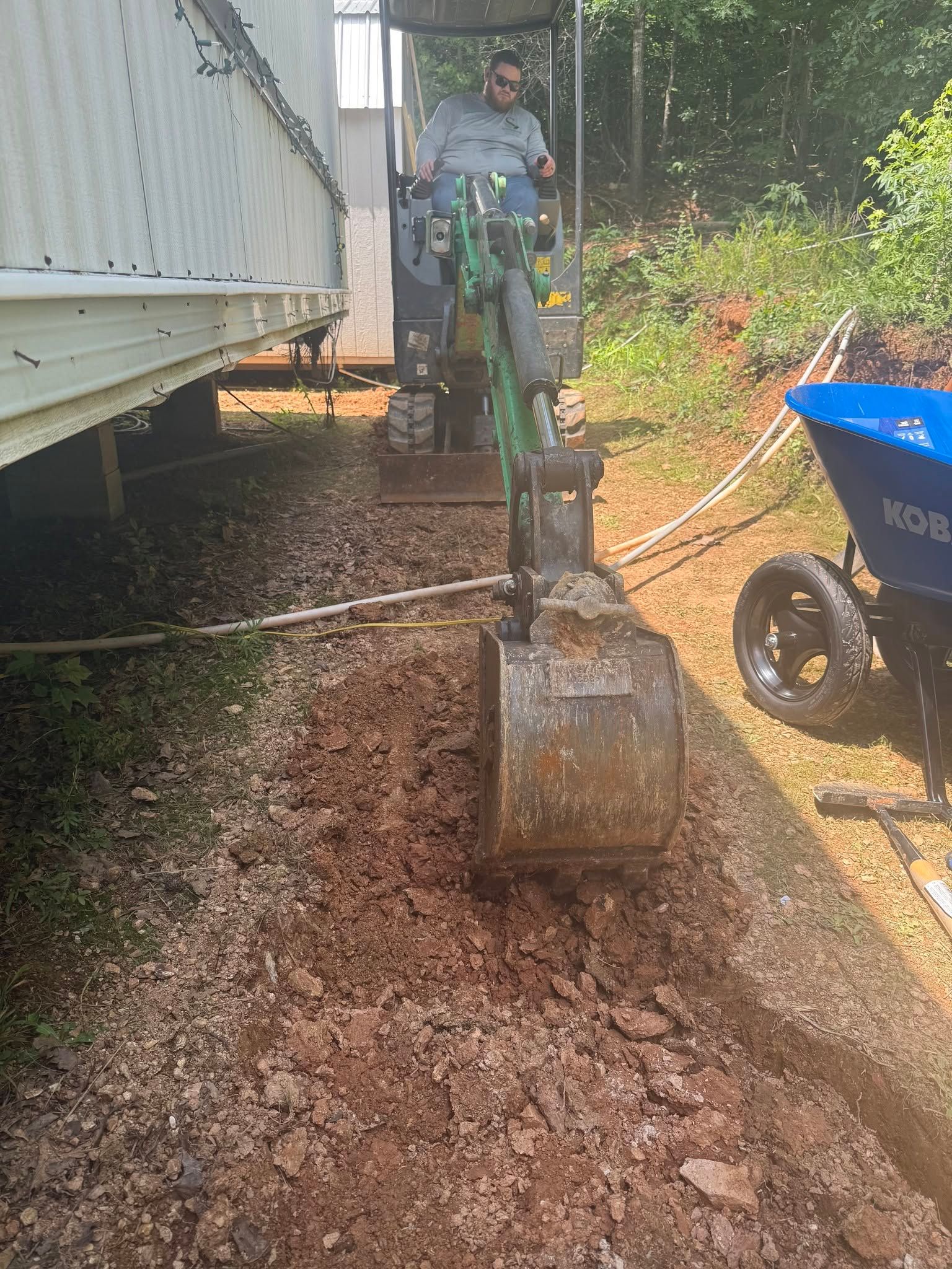 Person operating a green mini excavator beside a white trailer, digging gravel along a dirt path.