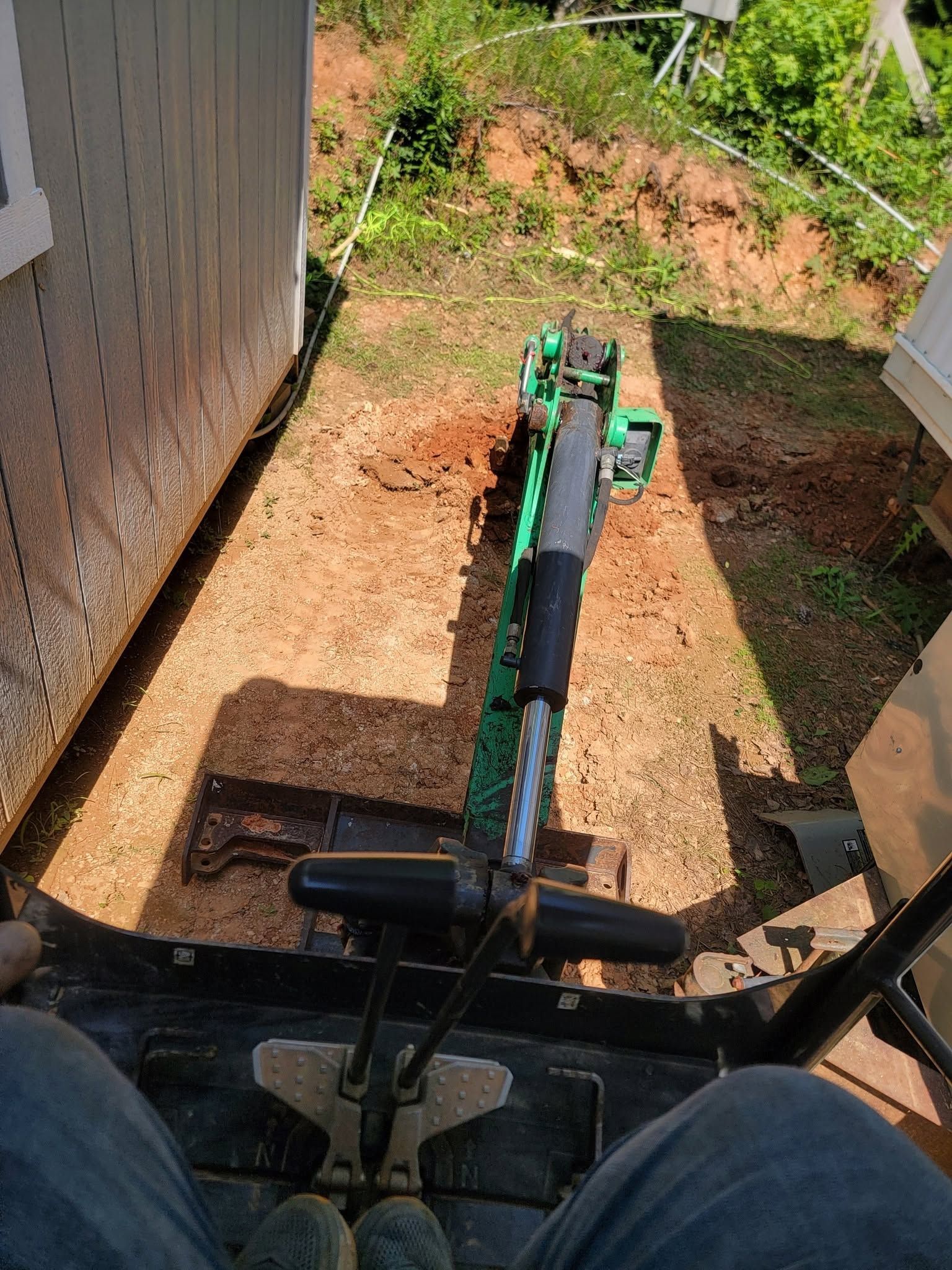 View from a tractor seat showing a green excavator arm working beside a fence in a dirt yard.