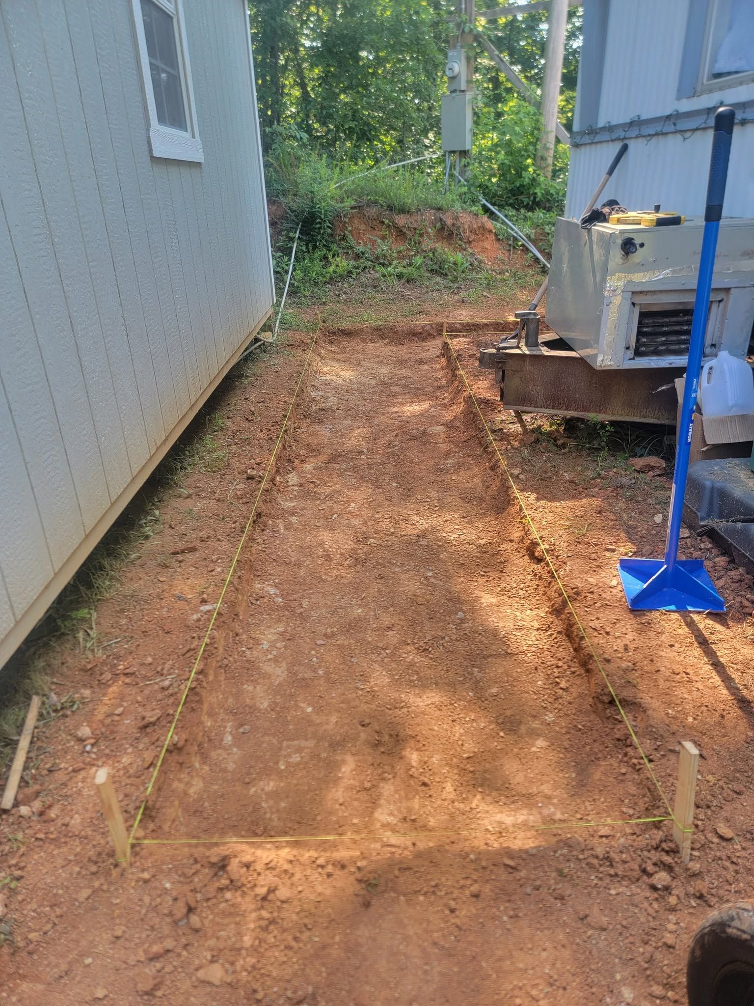 Narrow dirt path between a house and fence, with soil piled at the end and a blue post on the right.
