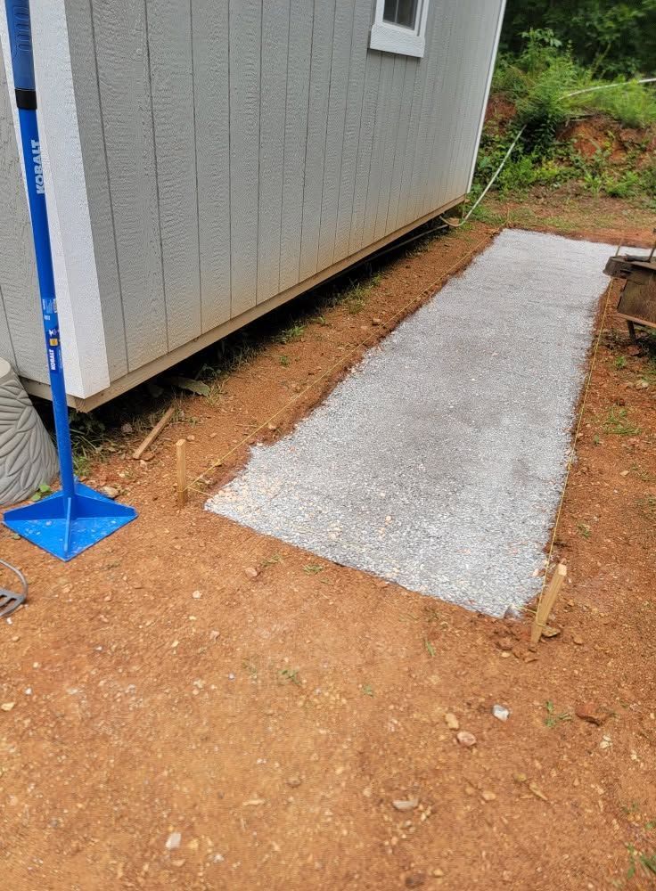 Concrete walkway beside a gray shed, with a blue broom and dirt path along the edge.