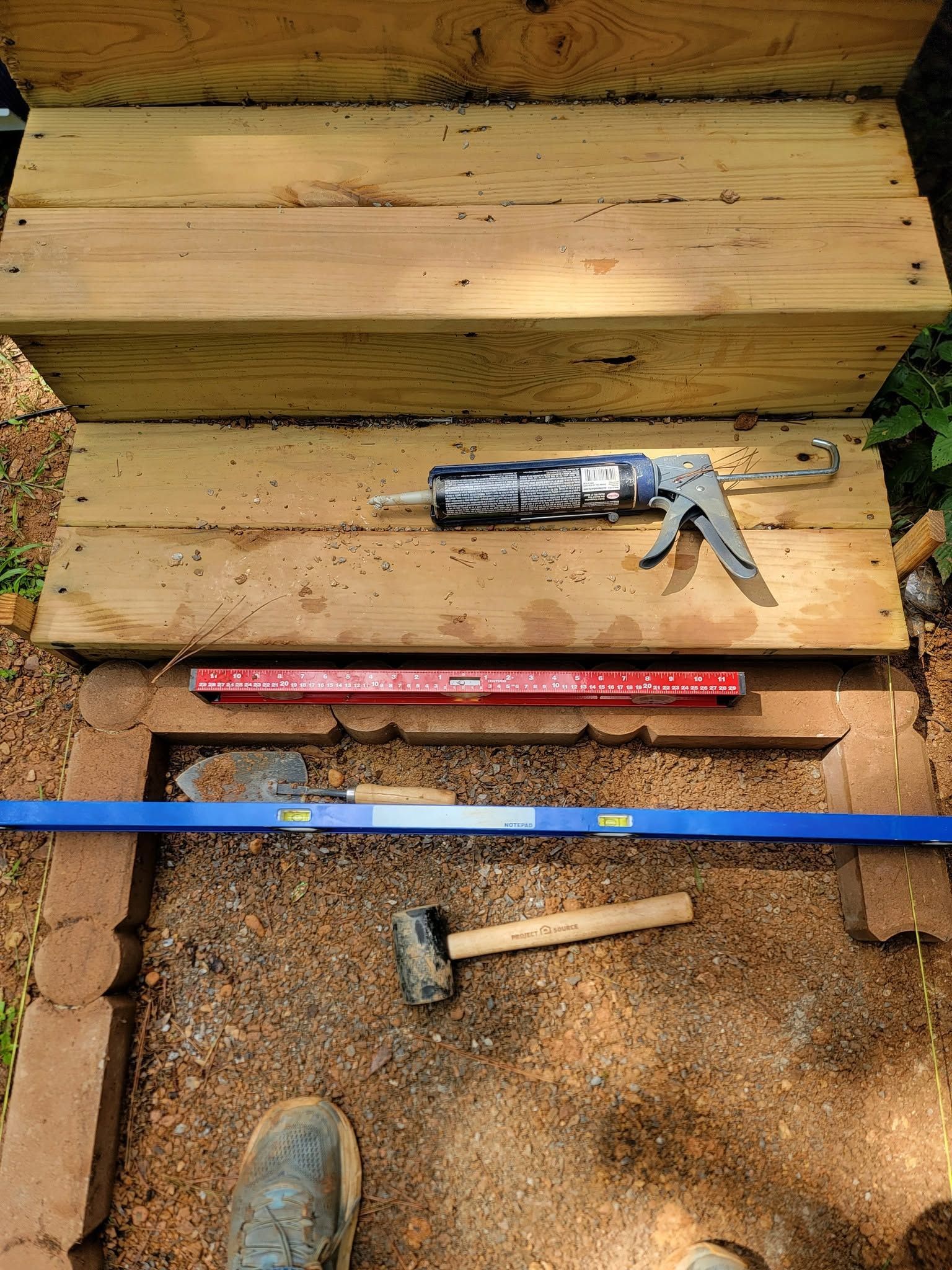 Tools on a wooden surface: a caulk gun, level, hammer, and boot near stairs with sawdust around them