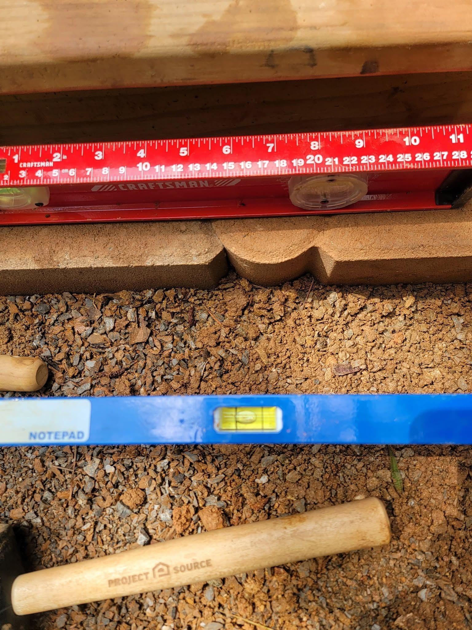 Blue and red level tools on a rough floor beside a wooden plank and tan handle object