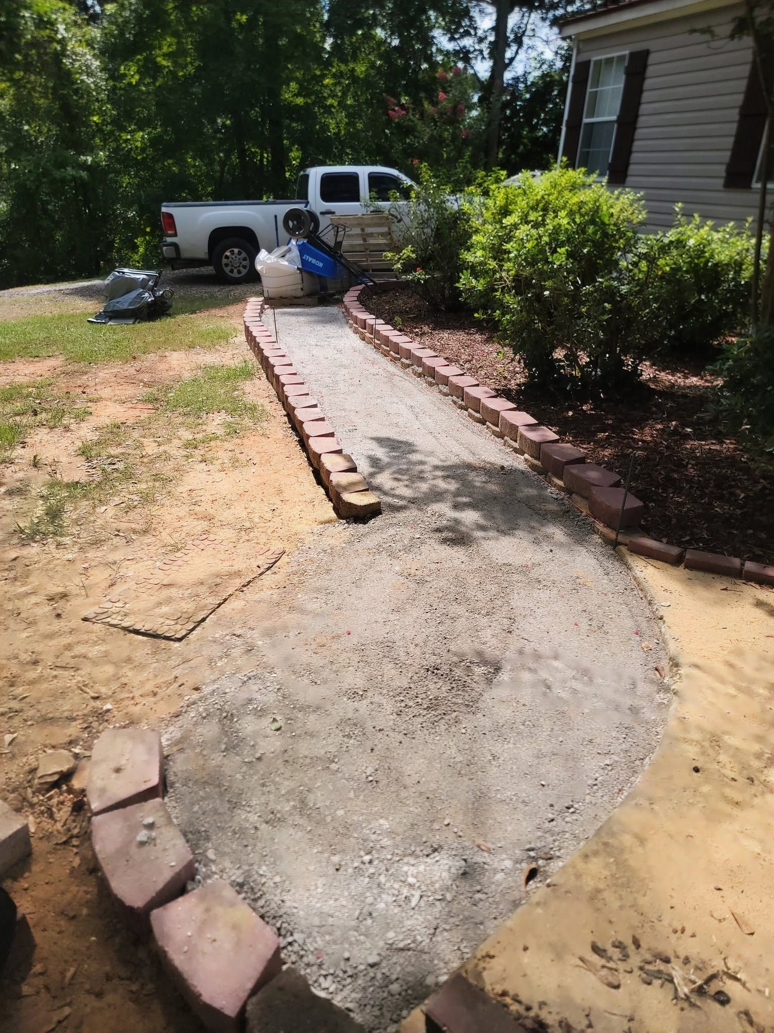 Partially finished concrete walkway beside a house, with bricks and a worker near a parked pickup truck.