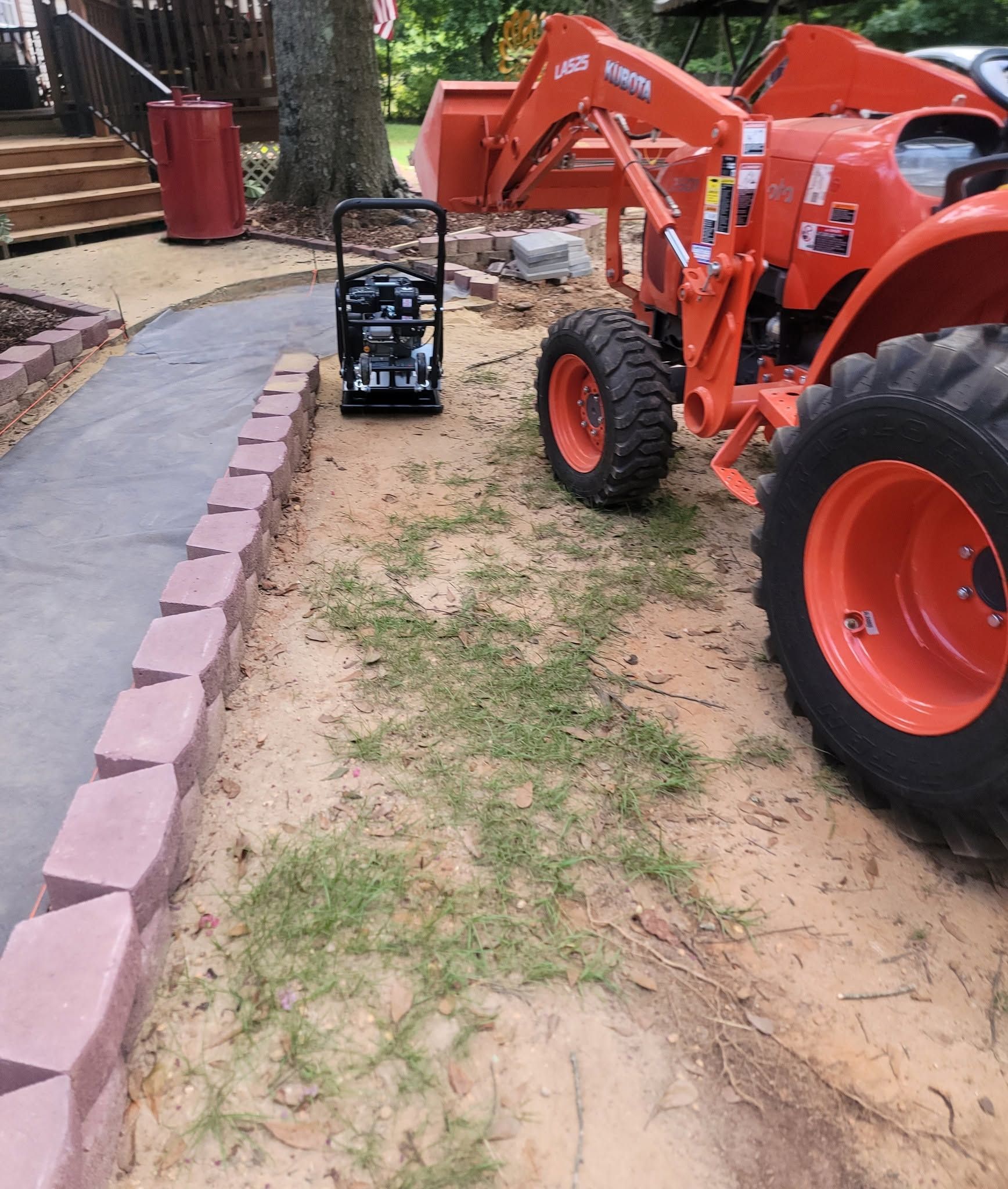 Orange tractor on a dirt path beside a pink curb near a parked stroller and trees