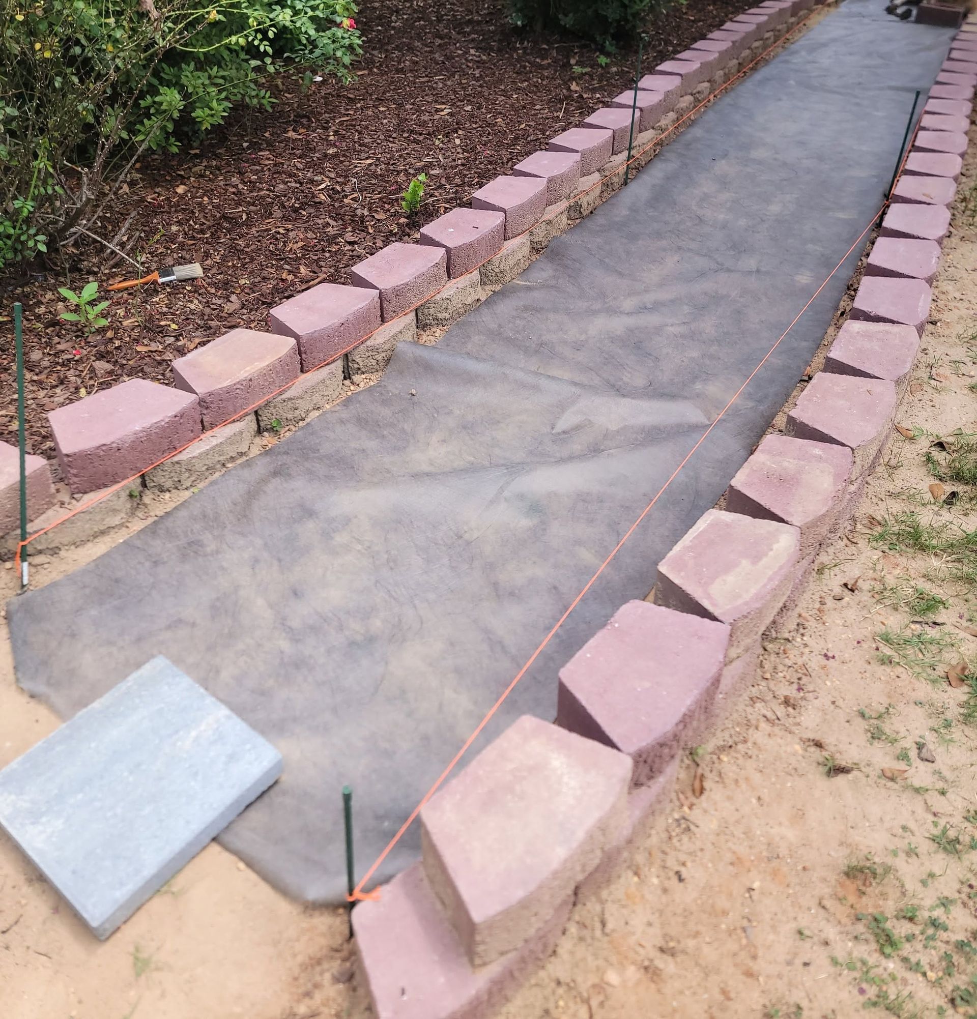 Concrete walkway under construction, bordered by pink bricks with a metal sheet ramp at the end.