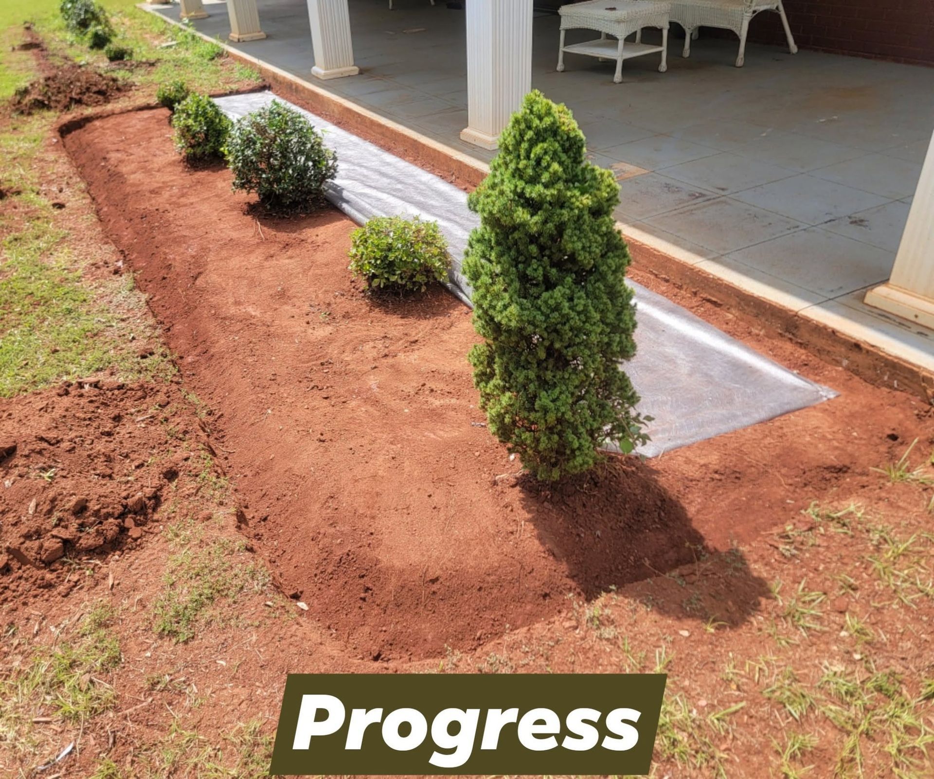 Freshly landscaped garden bed with red soil and small shrubs beside a porch, labeled “Progress”