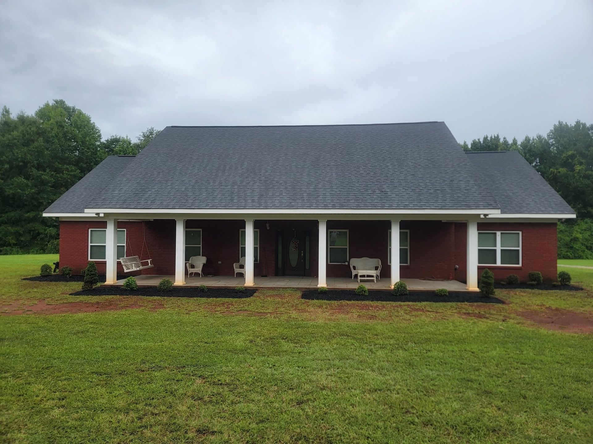 Single-story red brick house with a gray roof and white columns, set in a grassy yard under cloudy skies