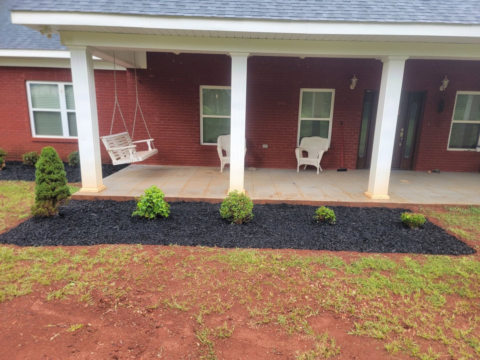 Covered porch with white columns, red siding, and white chairs behind a mulched garden bed with shrubs.