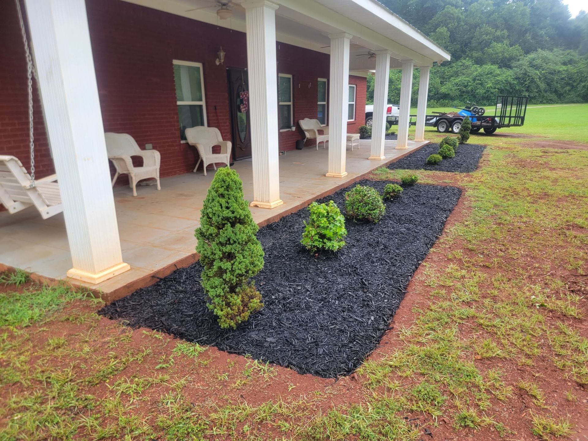 Red house porch with white columns and chairs beside a black-mulch landscape bed with small shrubs.