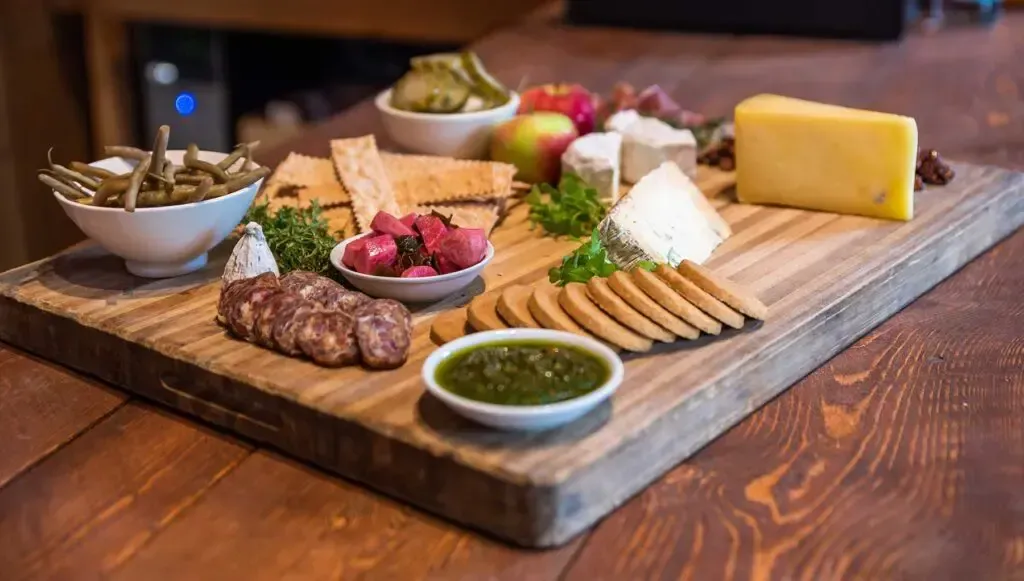 A wooden cutting board filled with cheese , crackers , fruit and vegetables on a wooden table.