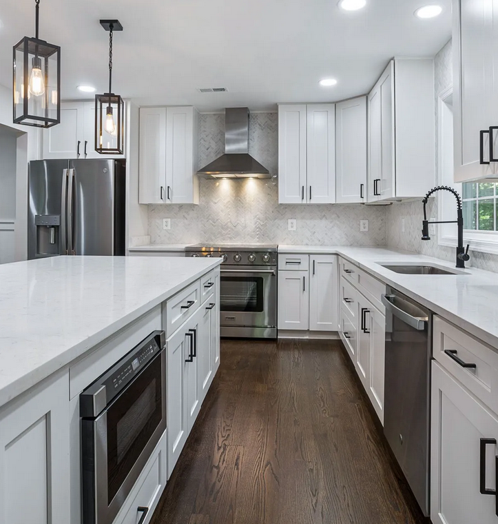 Modern white kitchen remodel with shaker cabinets, quartz countertops, stainless steel appliances, dark wood flooring, pendant lights, and a herringbone tile backsplash.
