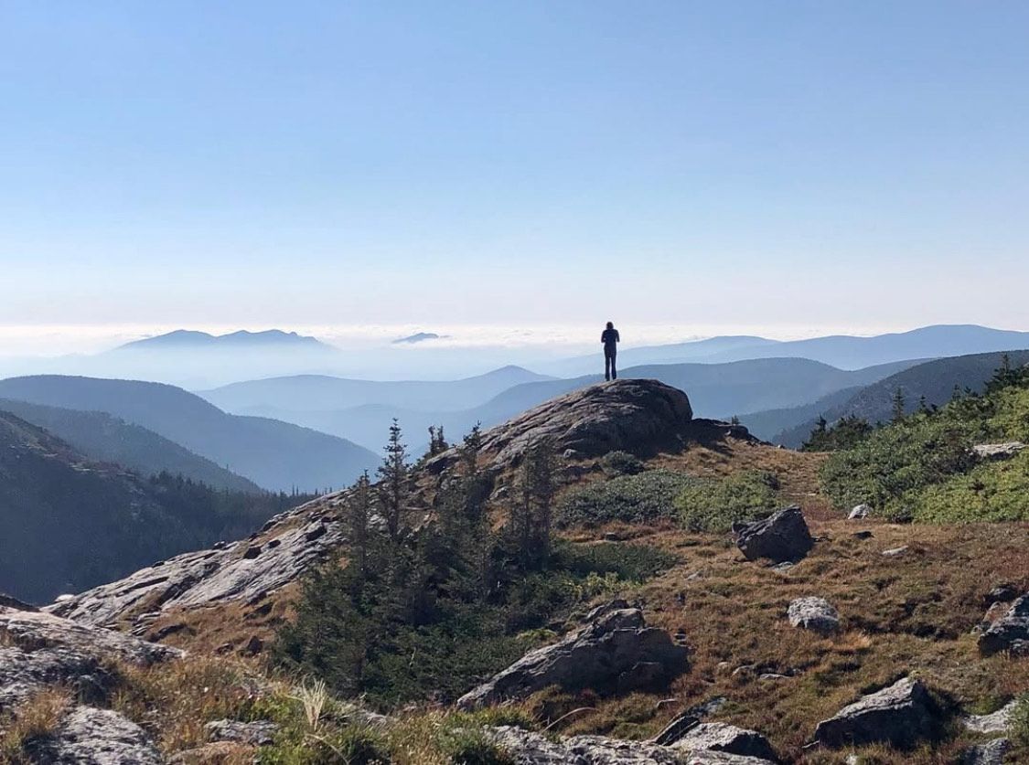 Hiker standing on a rocky mountain ridge overlooking layered blue valleys under a clear sky