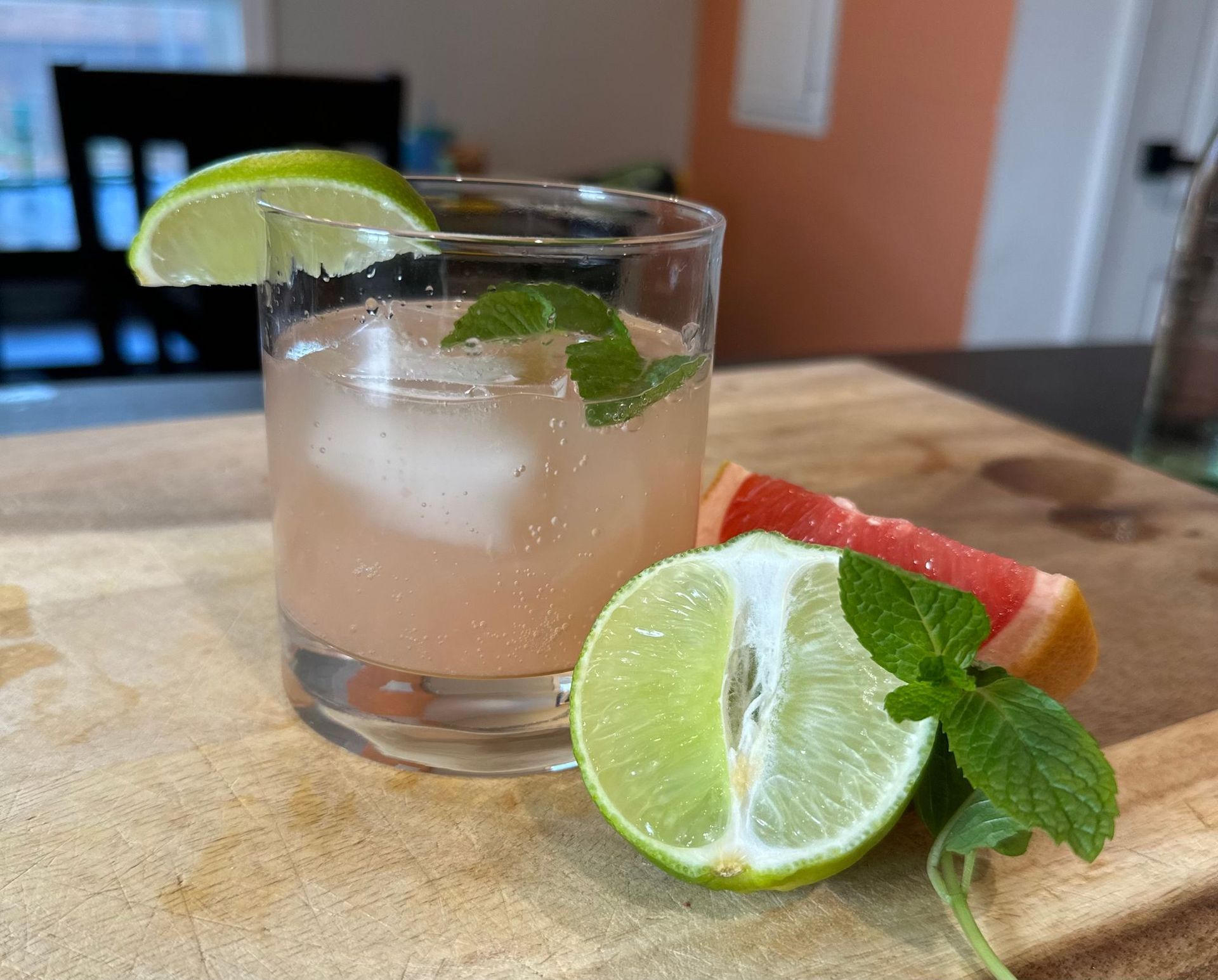 Pink cocktail in a rocks glass with lime and mint, beside a strawberry slice on a cutting board.