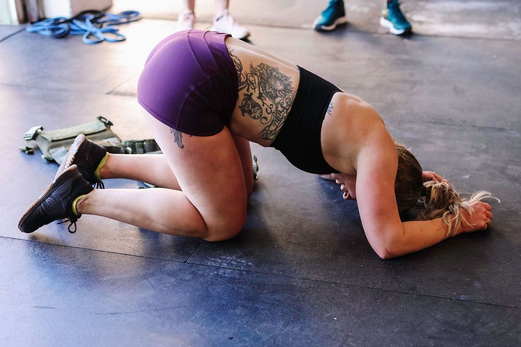 Person stretching on gym floor at Fortitude Fitness in a kneeling forearm pose, wearing black top and purple shorts.