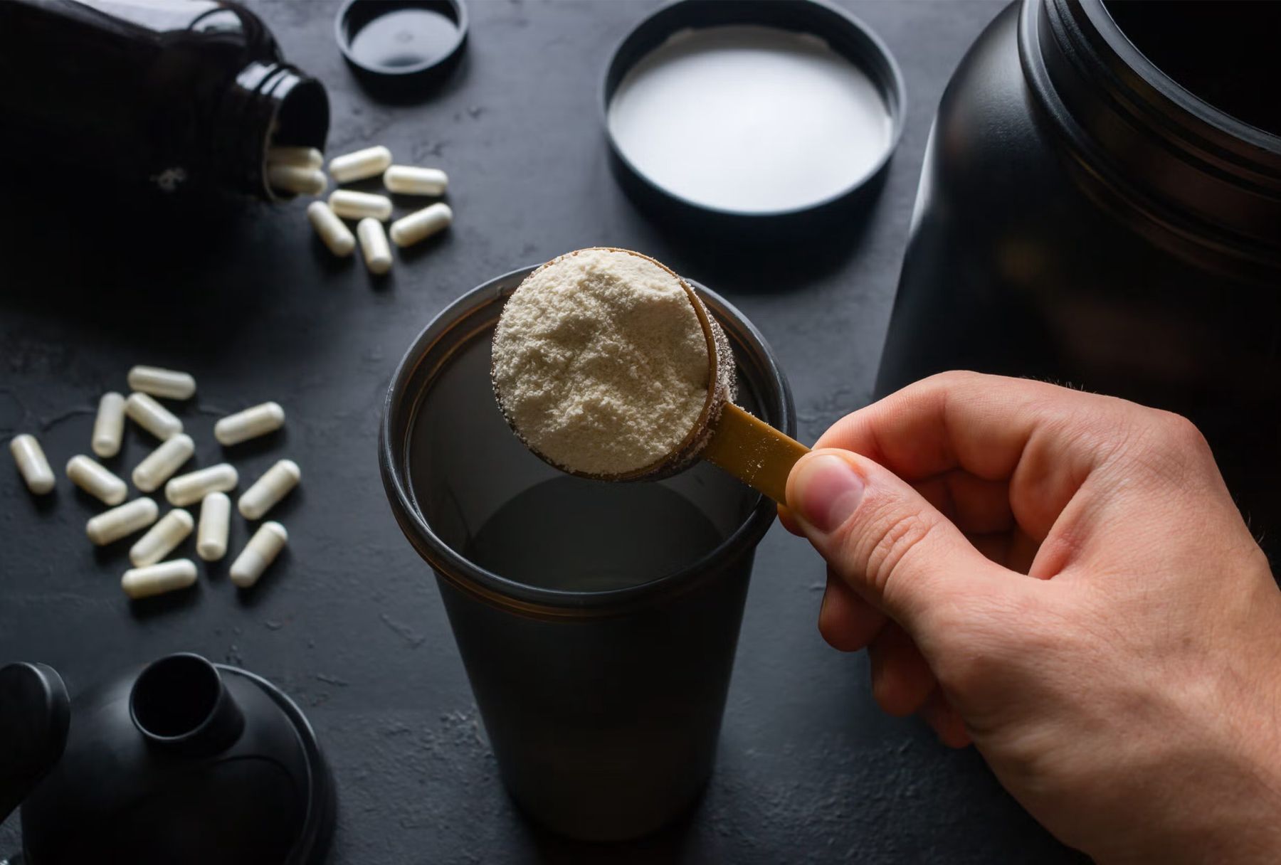 Hand pouring beige protein powder into a black shaker bottle beside capsules and a white drink