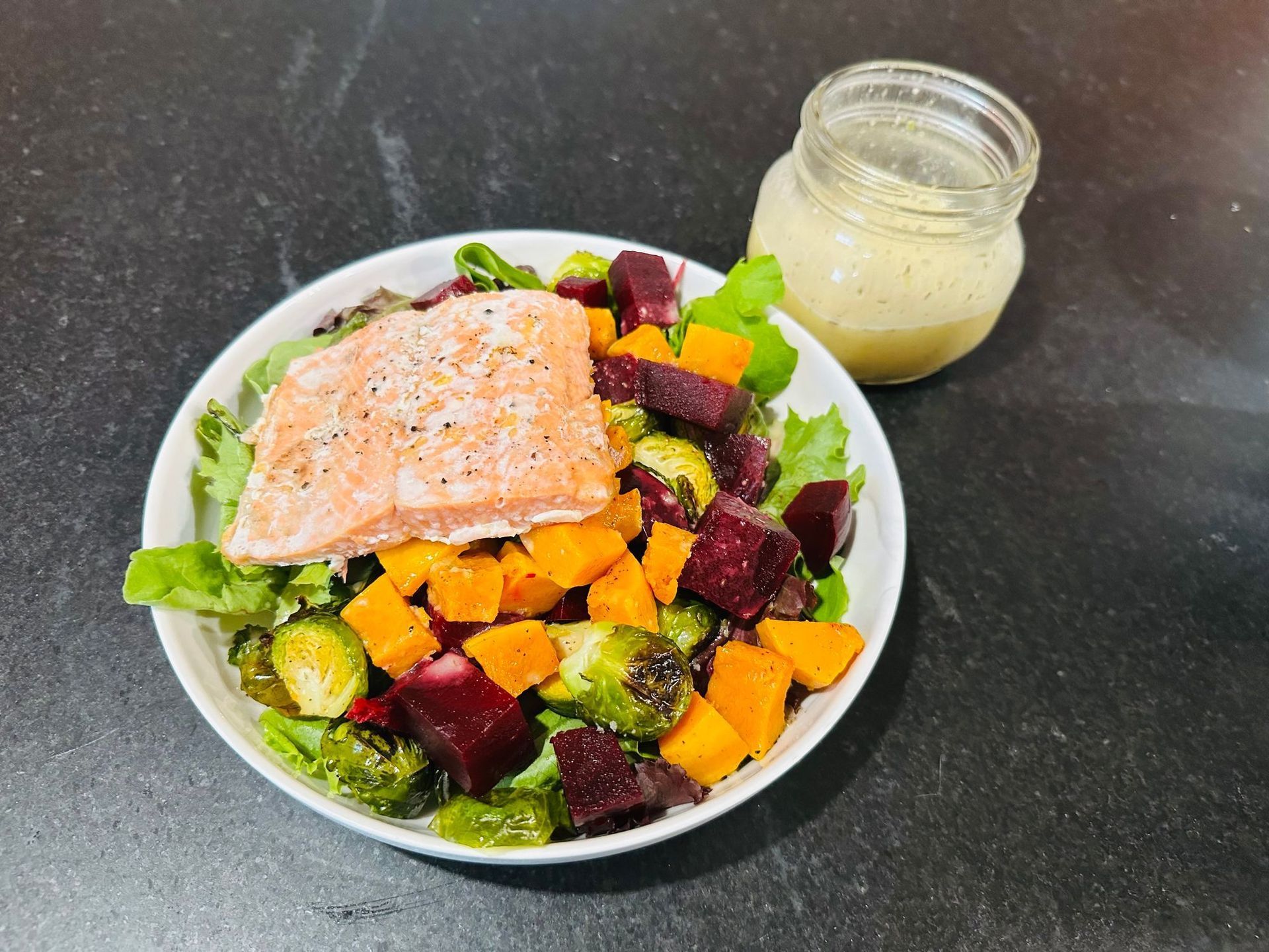Salmon salad with roasted vegetables in a bowl, with a jar of dressing on the side