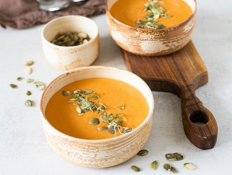 Two bowls of orange soup topped with seeds on a wooden board and a spoon, with a small cup nearby