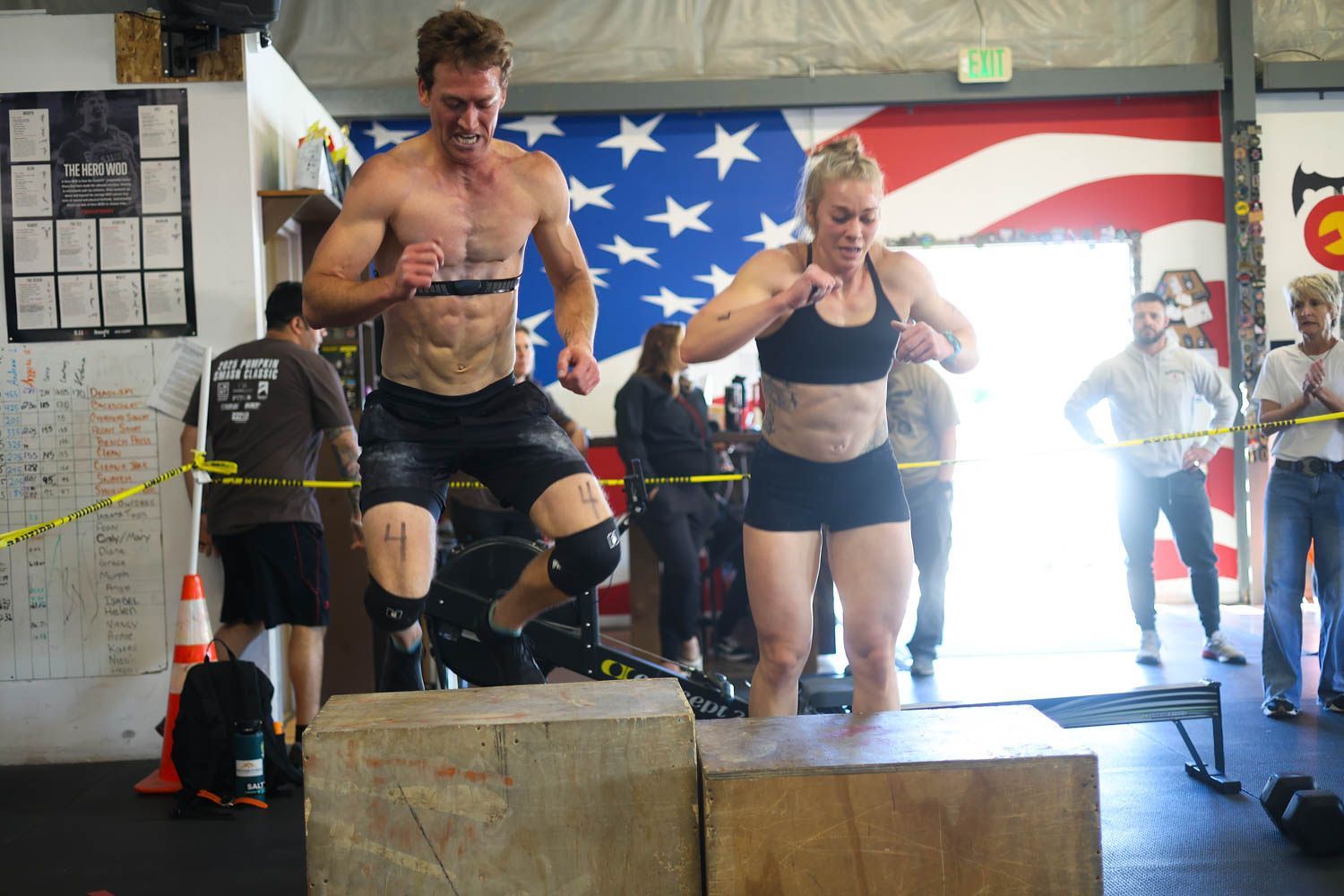 Two athletes jumping over wooden boxes in a competition area with an American flag backdrop at Fortitude Fitness