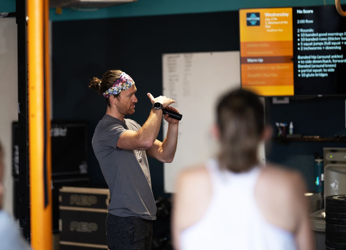 Man speaking into a microphone in a gym, with a blurred person in the foreground and a digital display behind him