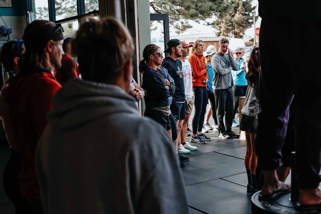People standing in a gym-like room near a speaker, gathered in a semicircle facing forward at Fortitude Fitness