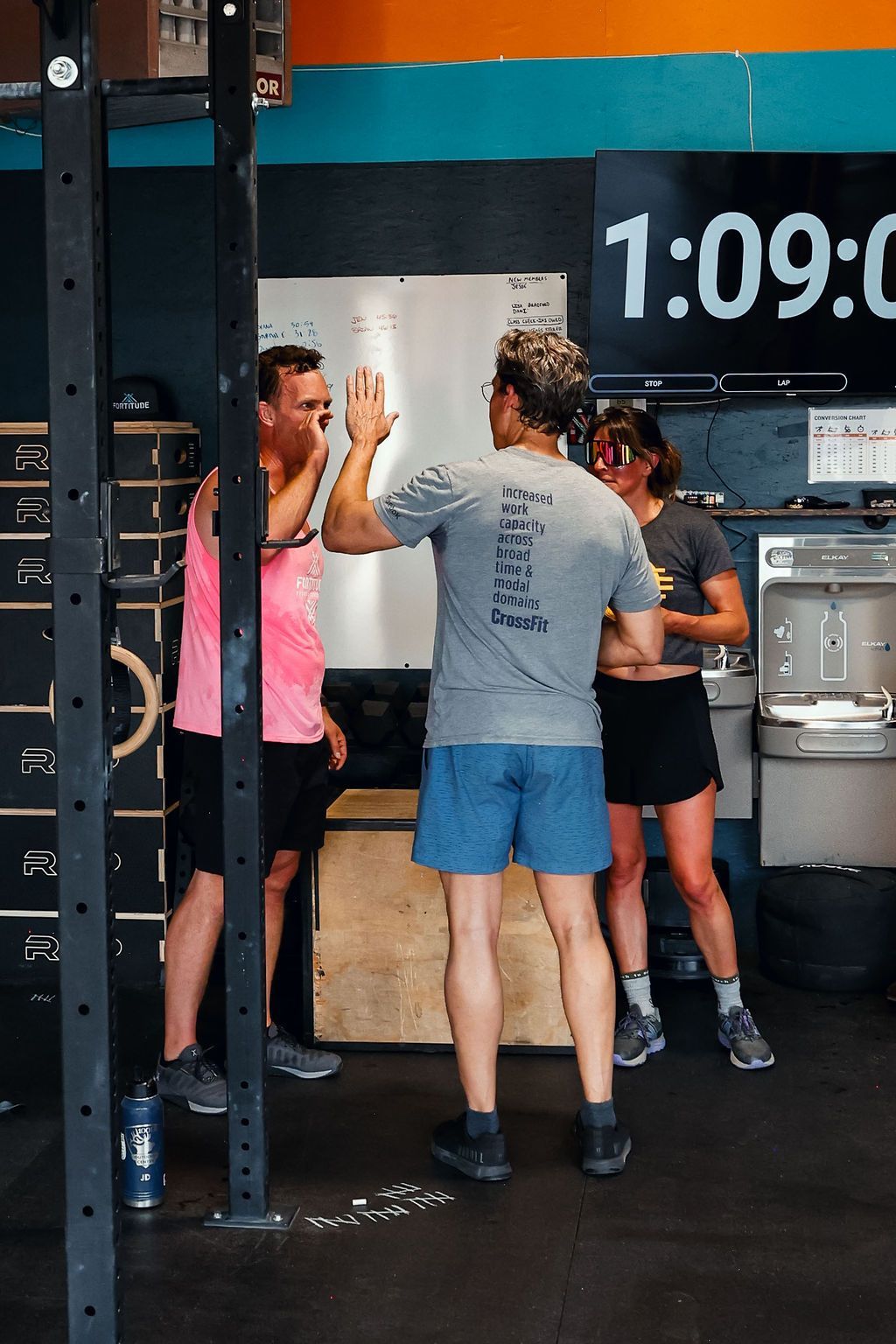 Three people high-five at Fortitude Fitness, with a wall clock and equipment in the background.