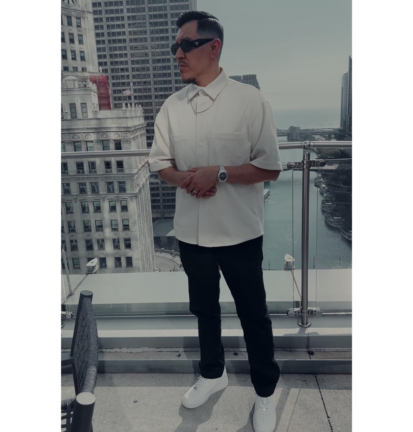 A man wearing sunglasses and a white shirt is standing on a balcony overlooking a city.