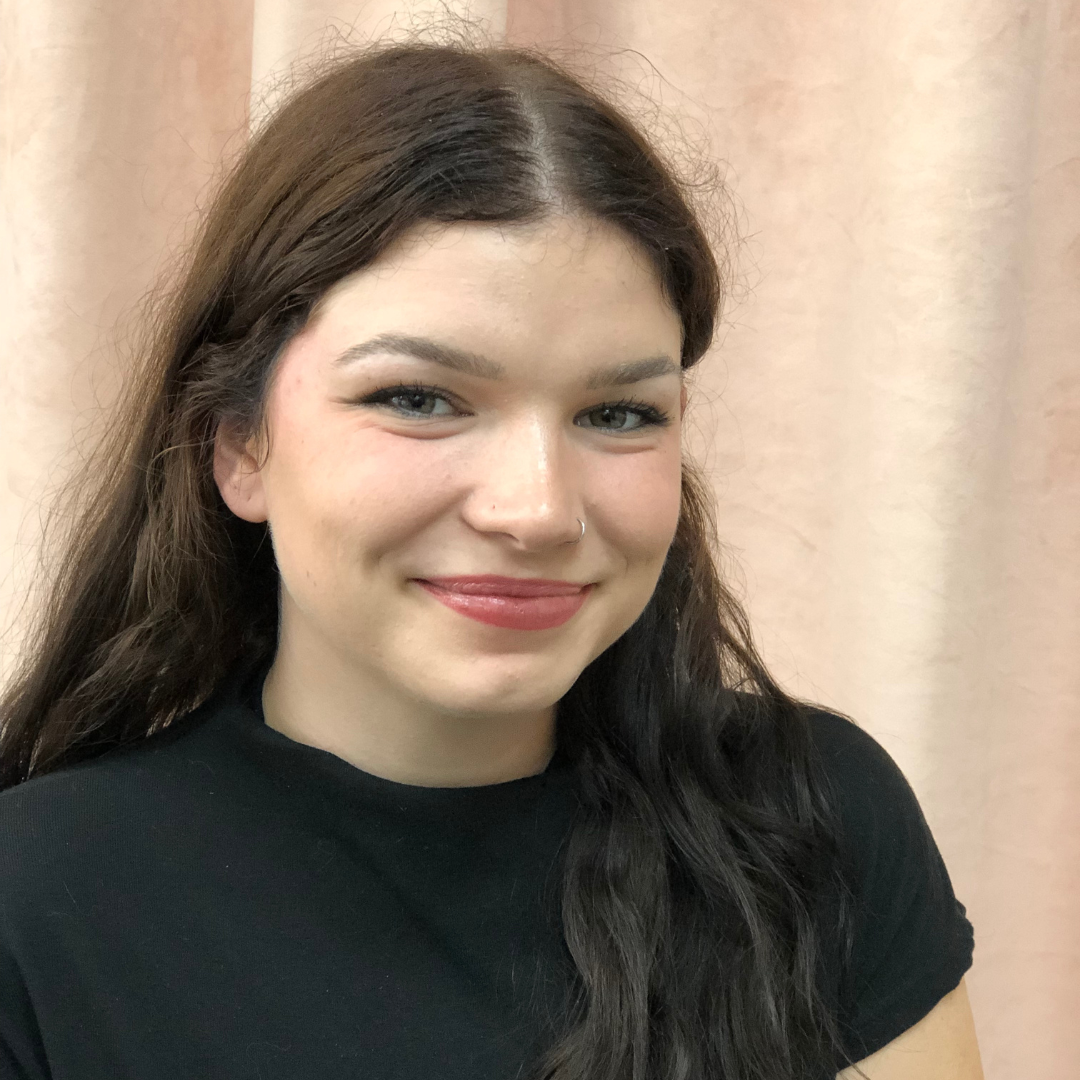 Une femme aux cheveux noirs sourit, vêtue d'une chemise noire, sur fond rose.