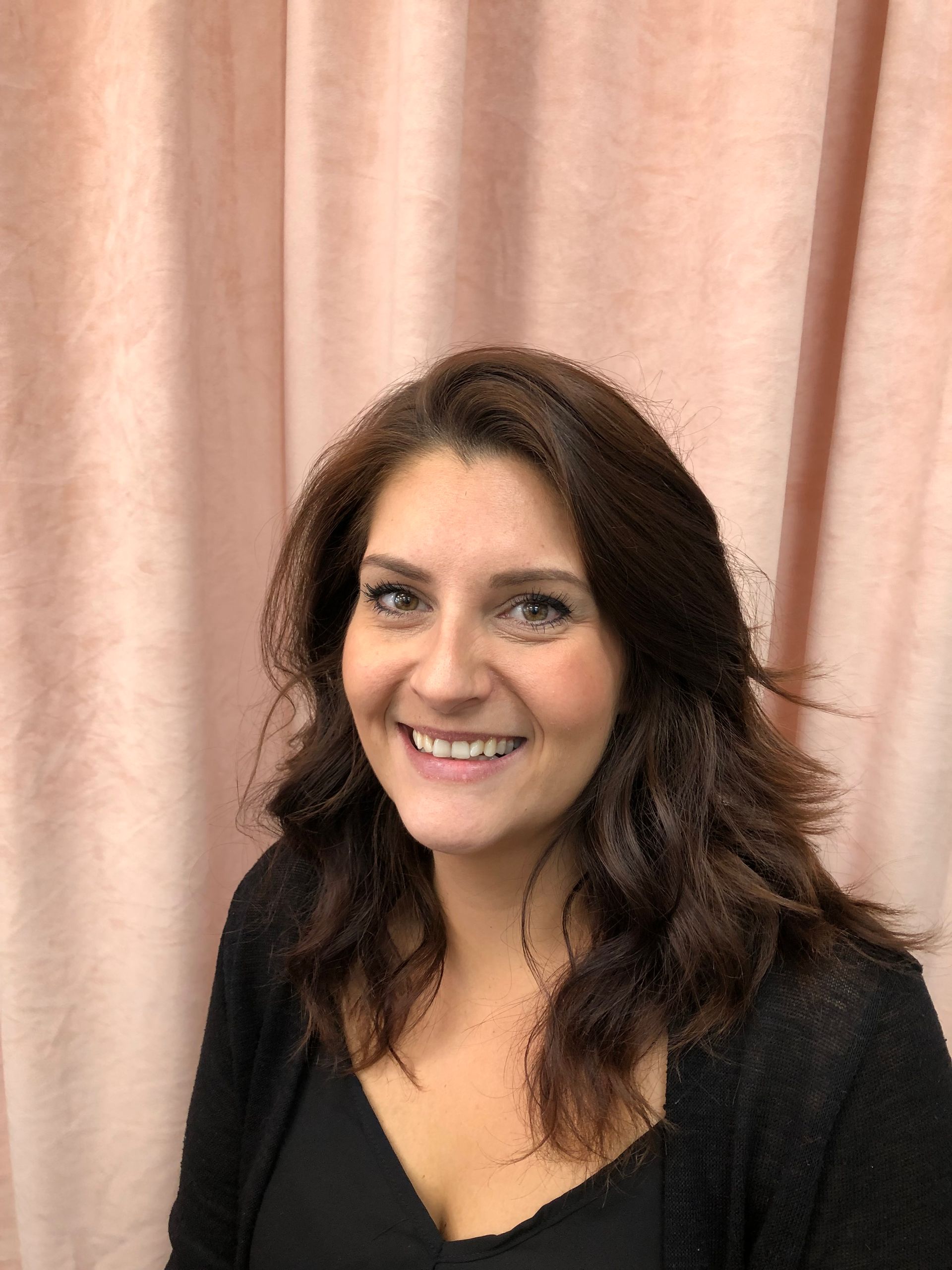 Femme aux cheveux bruns souriant devant des rideaux roses.
