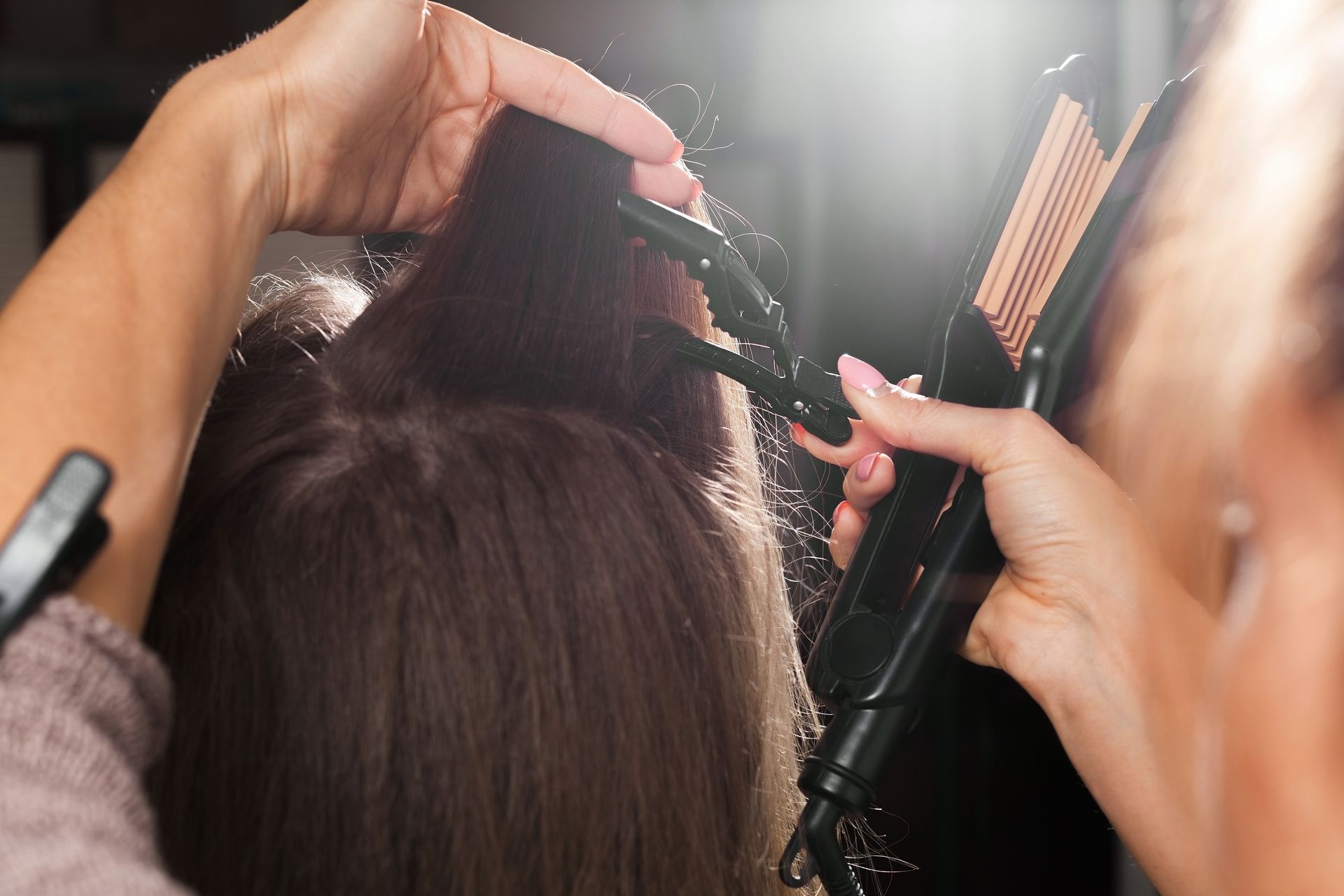 Mains utilisant une pince à friser et un fer plat sur des cheveux bruns dans un salon.