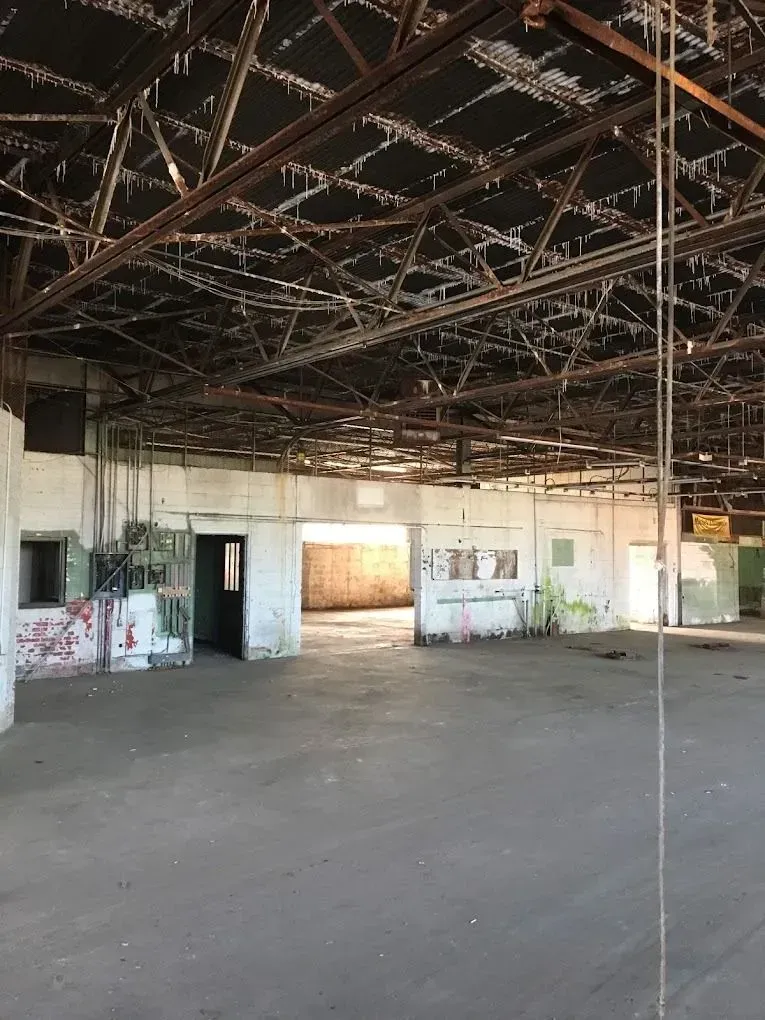 Interior view of a large, dilapidated warehouse. Concrete floor, metal roof structure, and open doorways.