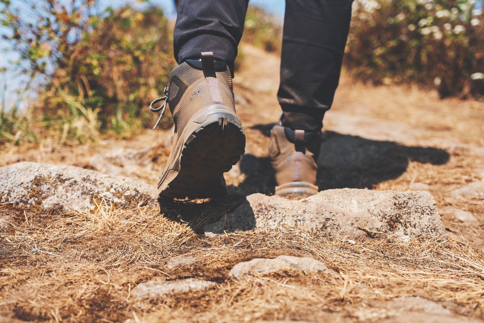 Booted feet walking on a rocky dirt trail in a sunny outdoor setting