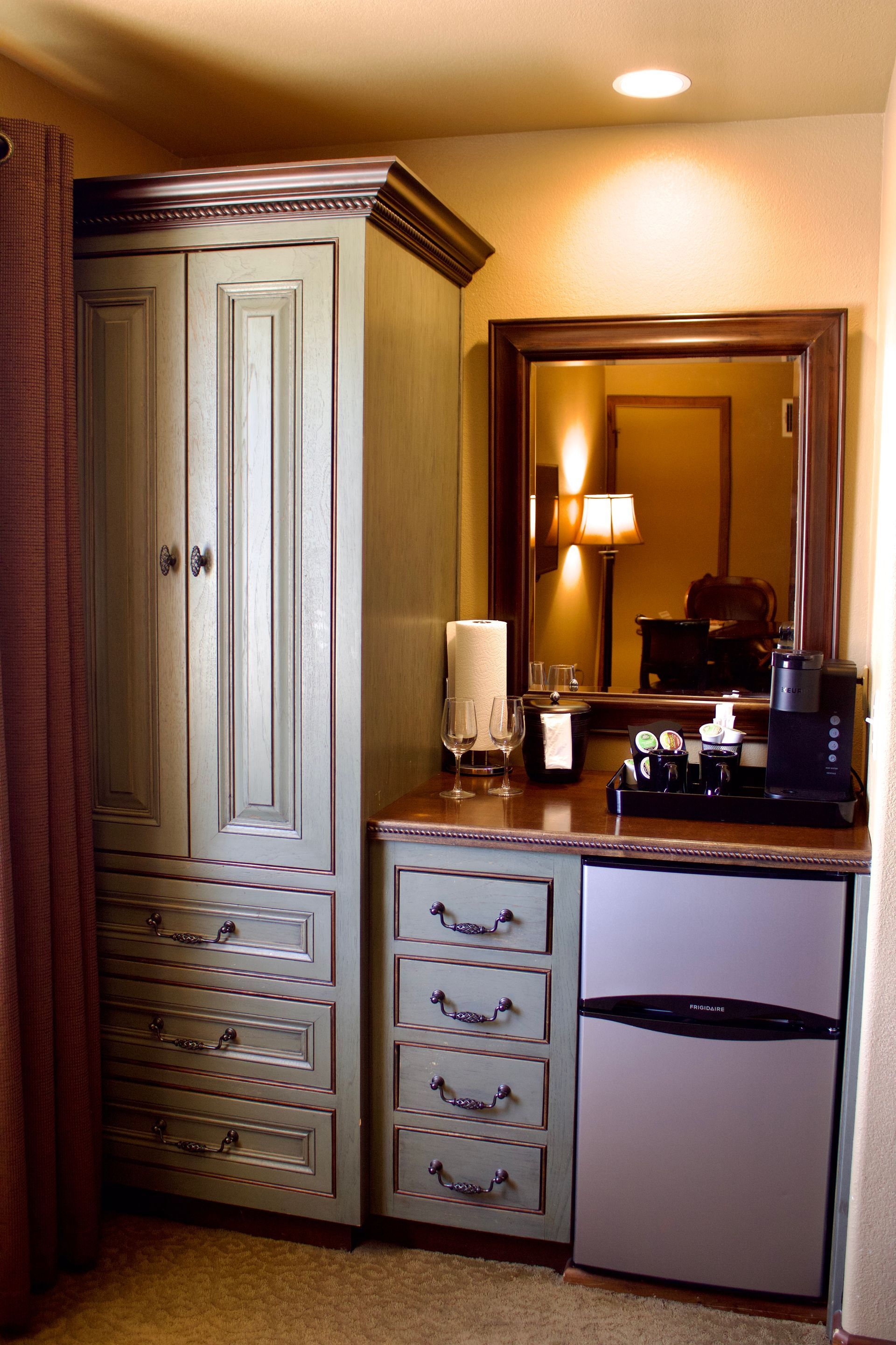 Hotel vanity with white armoire, drawers, mini fridge, mirror, and lamp on a dark countertop