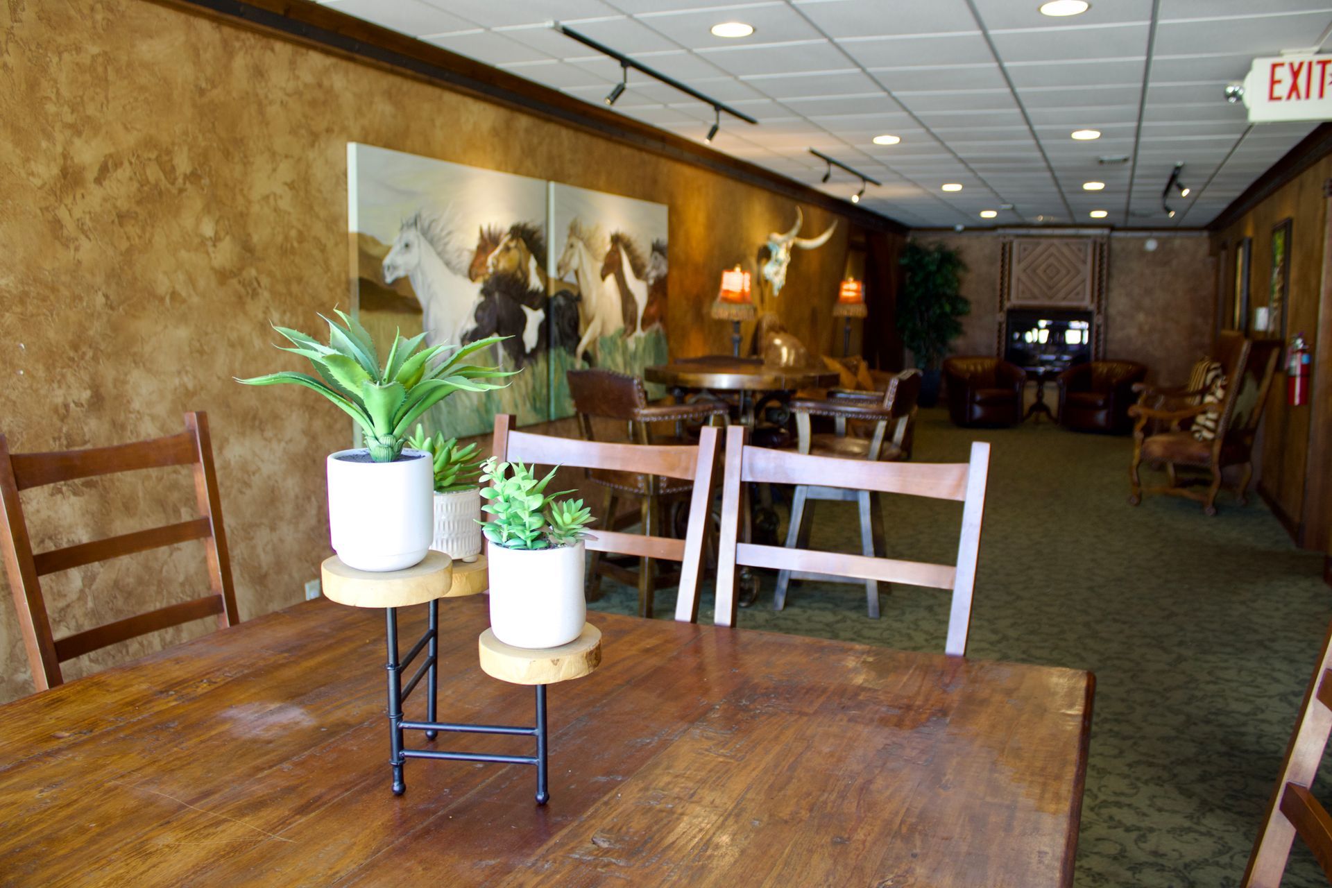Empty hotel hallway with tables, potted plants, wall art, and an exit sign at the far end