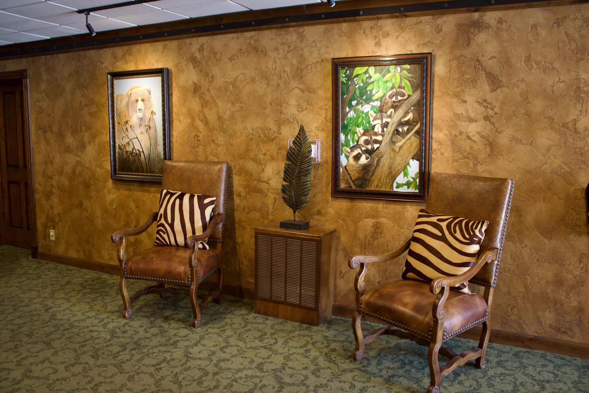 Lobby seating area with two wooden chairs, a small table, and framed wall art on a textured brown wall.