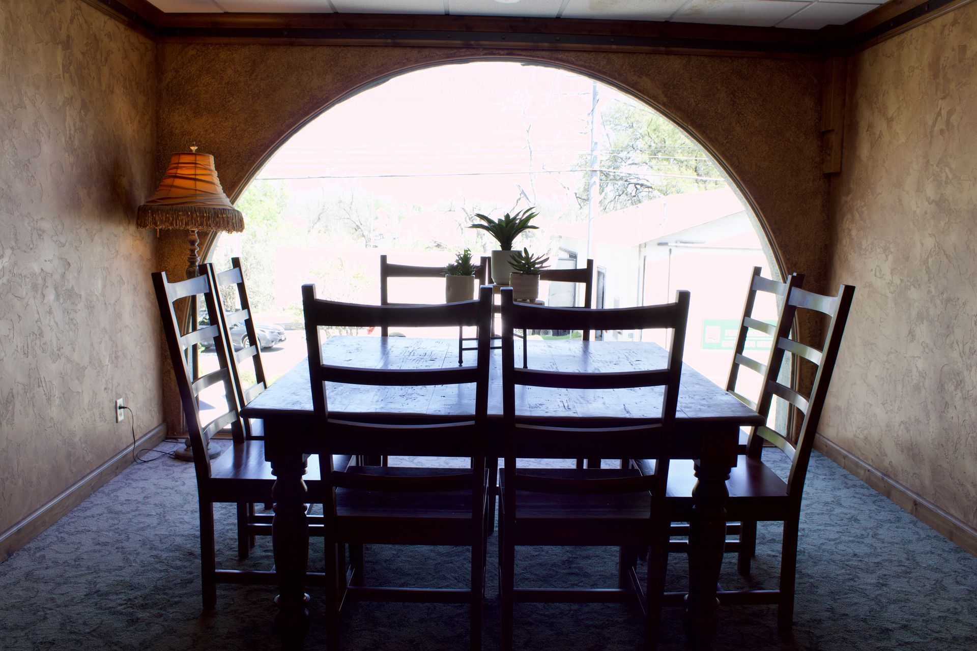 Dining table and chairs in a rustic room, with a large arched window and bright outdoor view.