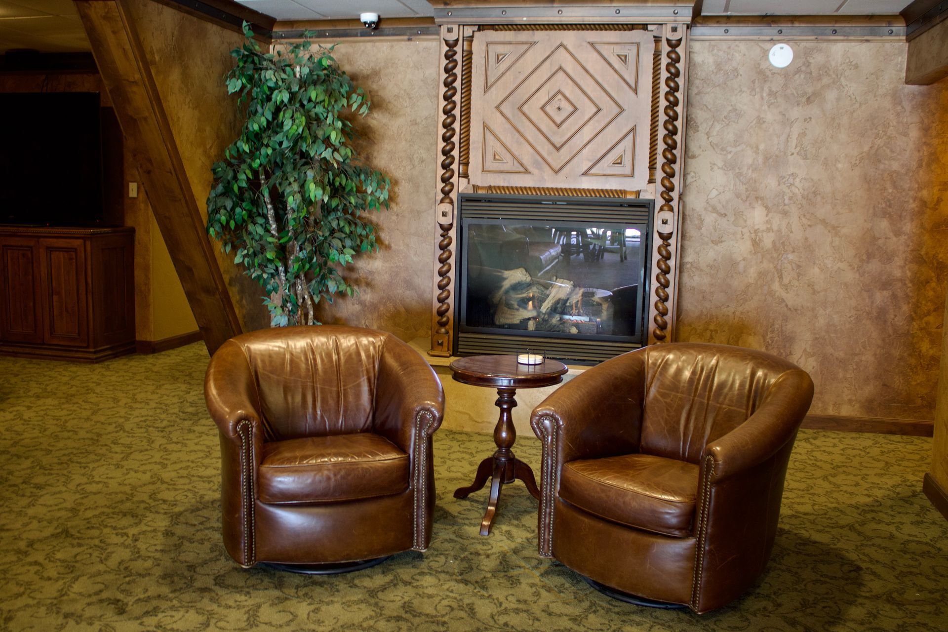Two brown leather chairs facing a small table in a carpeted lobby with a fireplace and potted plant.