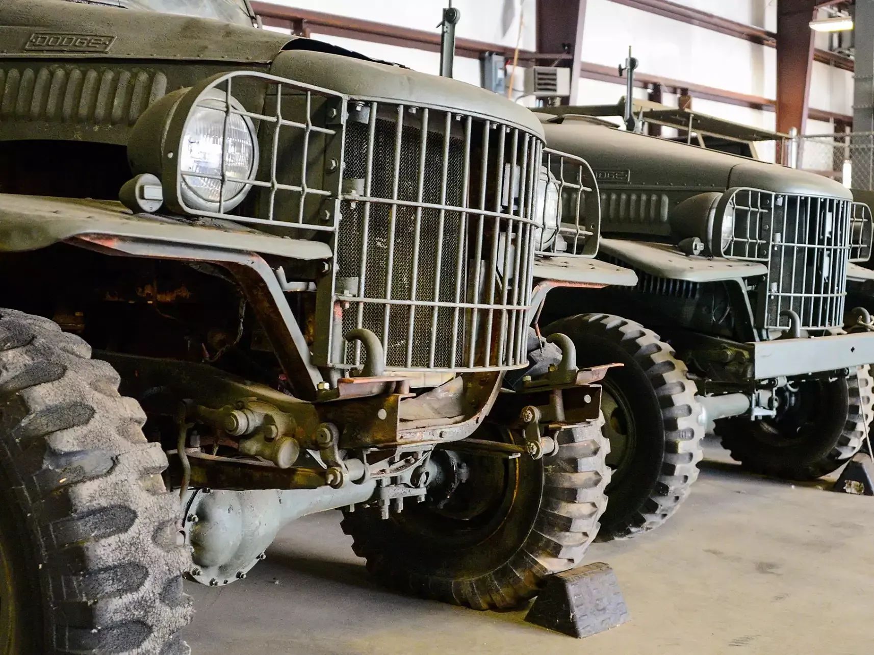 Two olive drab military trucks in a garage, showing large tires and front grilles.
