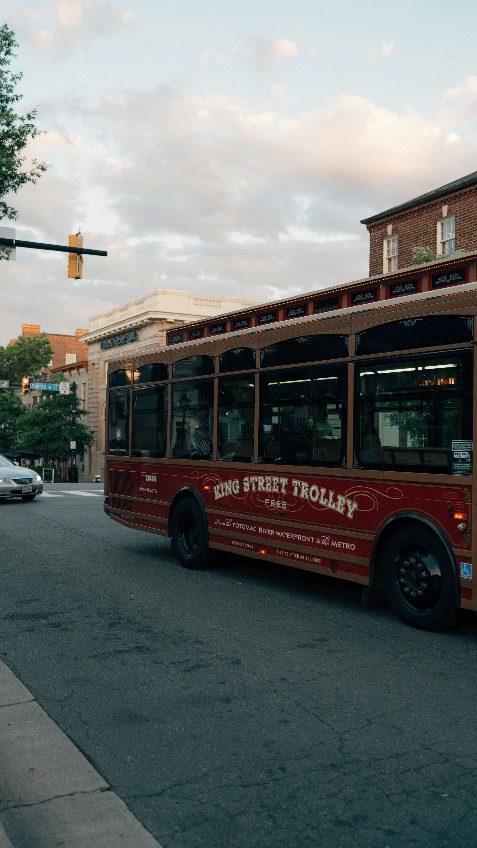Red trolley bus on a city street at dusk, with brick buildings and a traffic light nearby.