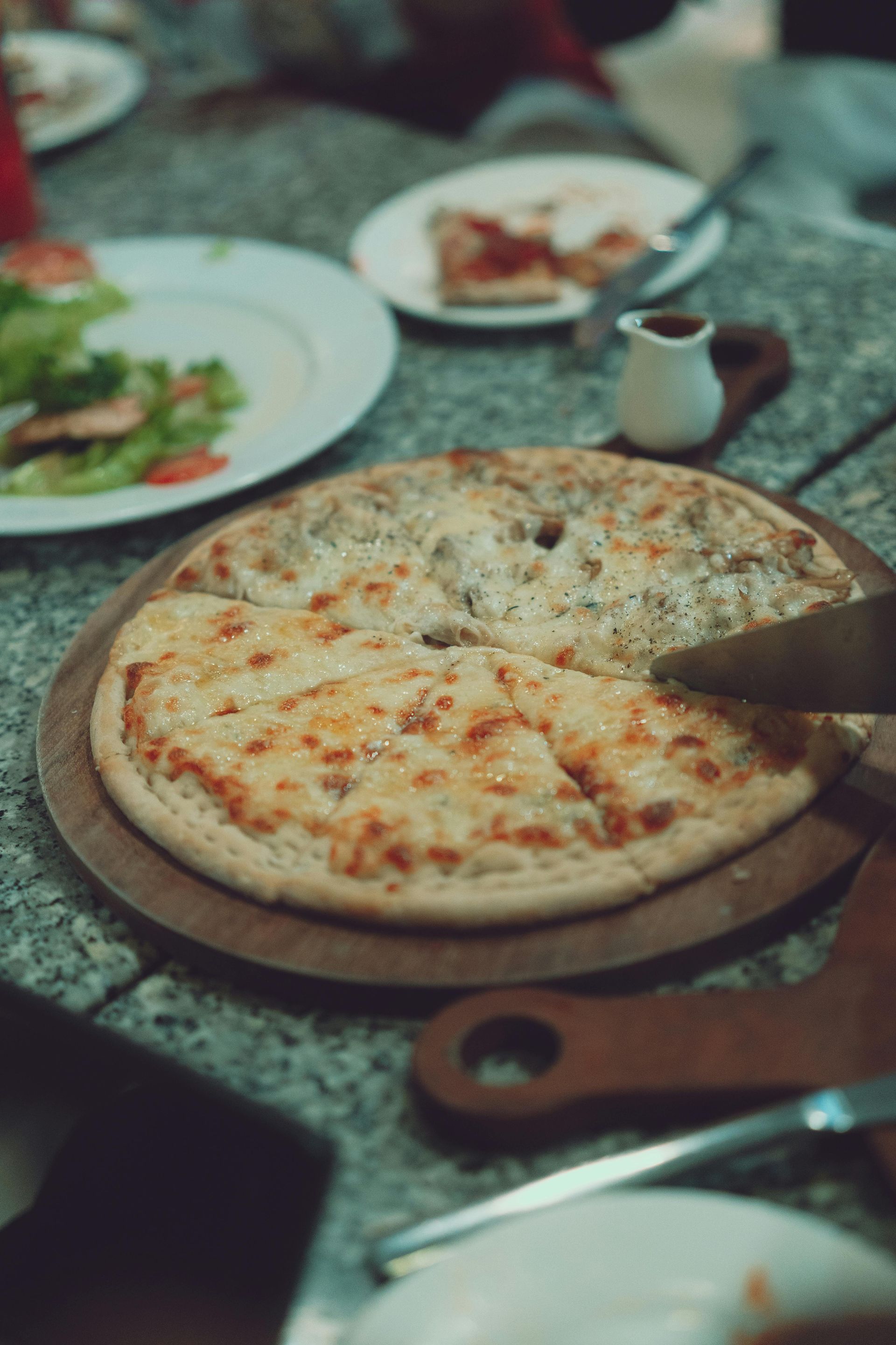 Pizza on a wooden board at a dining table, with other dishes and utensils nearby