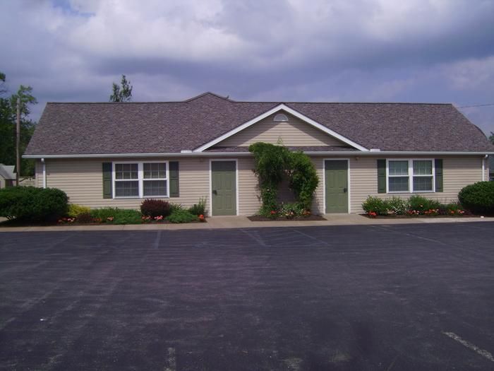 Tan, single-story building with two green doors and windows, dark roof, surrounded by a parking lot and small bushes.