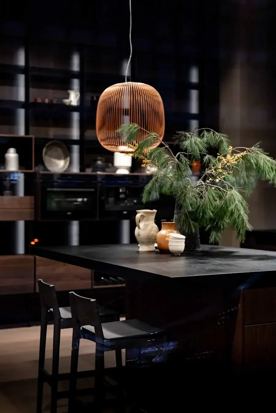 Modern kitchen with dark cabinets, island with greenery, and a hanging wooden light fixture.