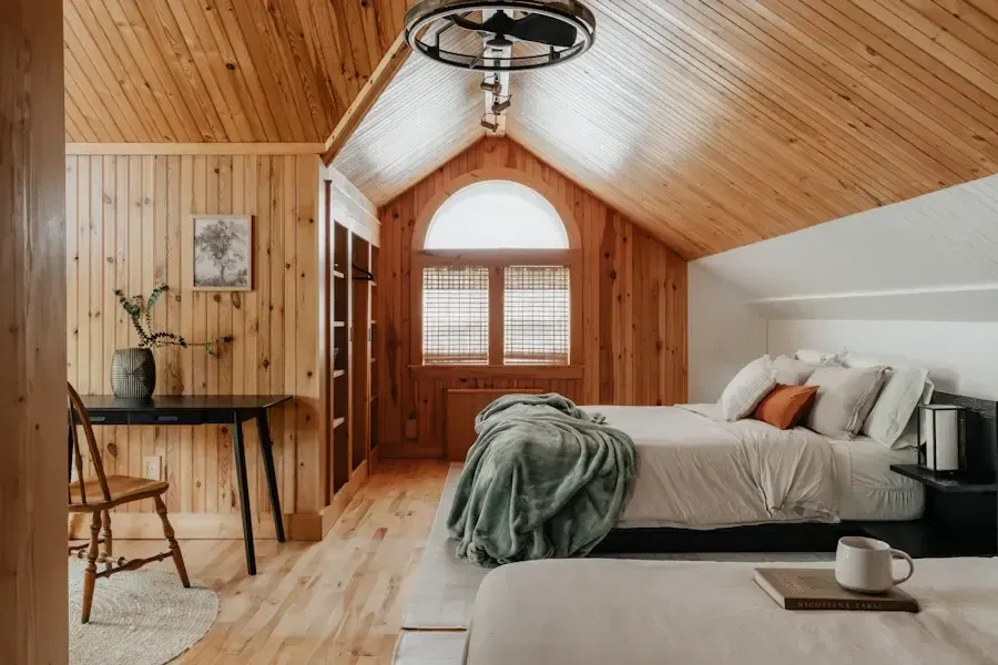 Bedroom with wood-paneled walls, two beds, desk, arched window, and ceiling fan.